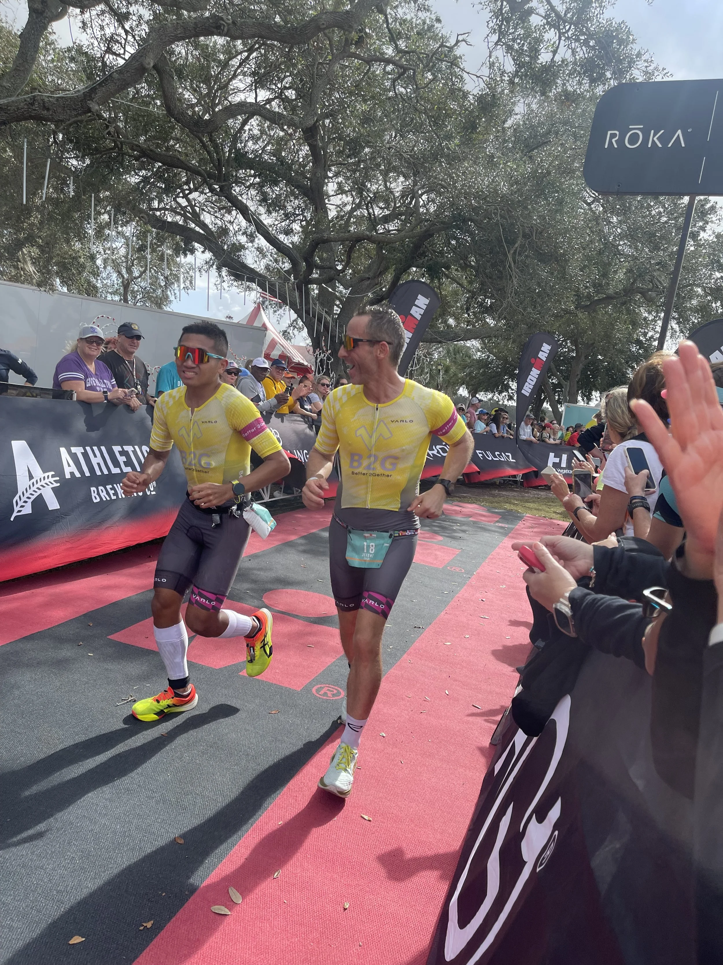 Two male athletes in yellow and black running uniforms smiling and celebrating as they finish a race, with spectators cheering on the sides under large trees and event flags in the background.