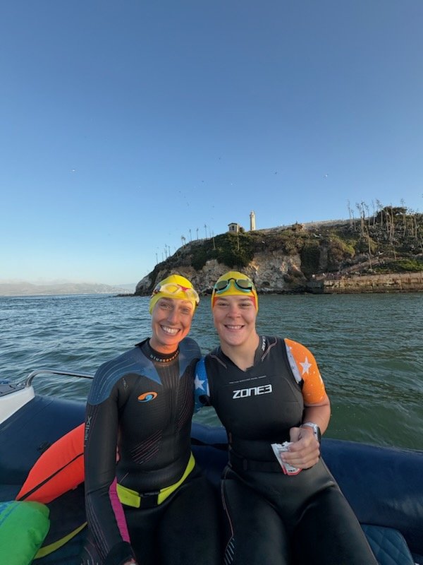 Two women in wetsuits and yellow swim caps sitting on a boat with the water and a rocky shoreline with a lighthouse in the background.