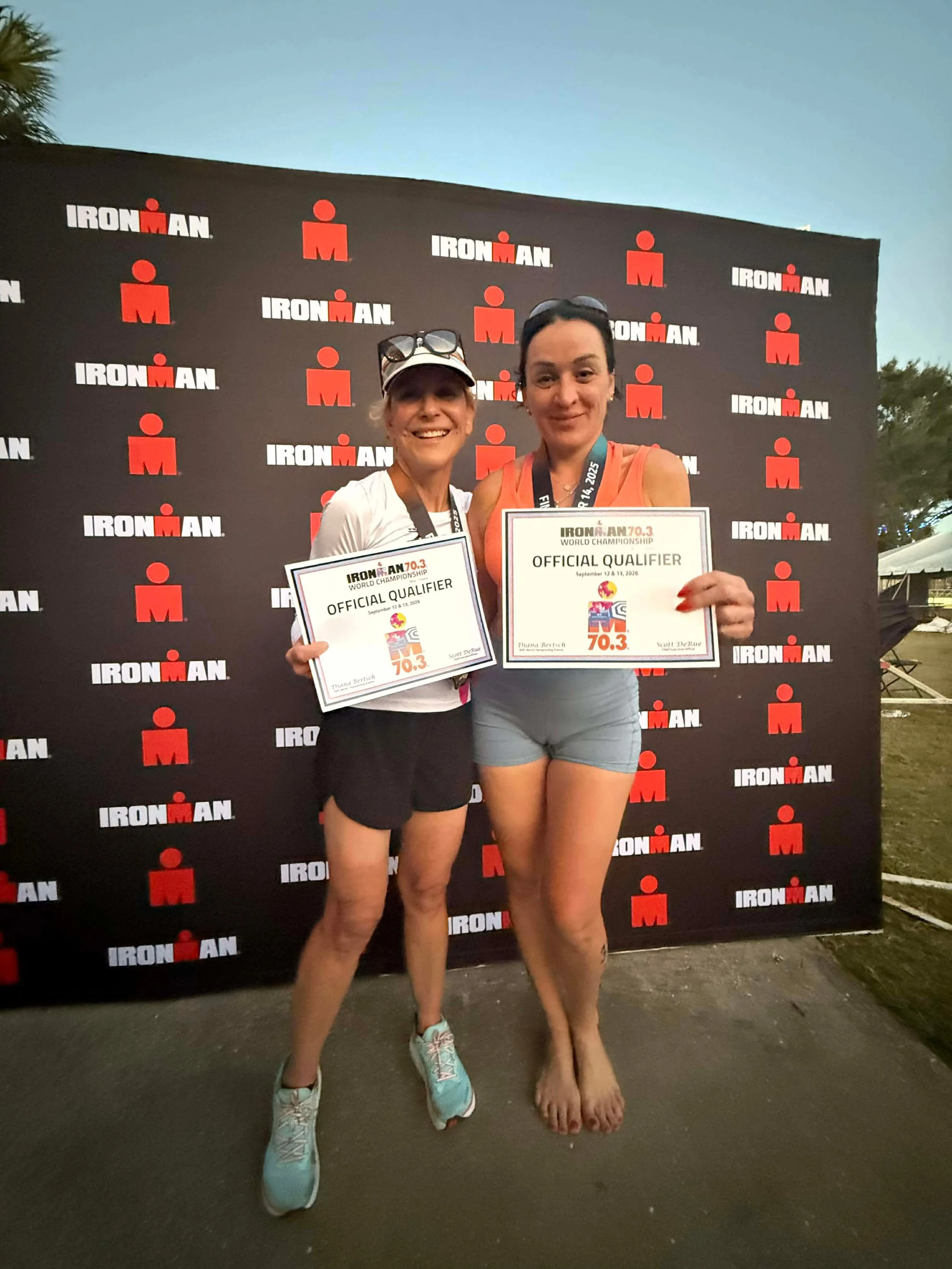Two women standing in front of an IRONMAN event backdrop, holding certificates that say 'Official Qualifier.' One woman is wearing a white shirt, shorts, and running shoes, and the other is in a tank top and shorts, holding her certificate up.