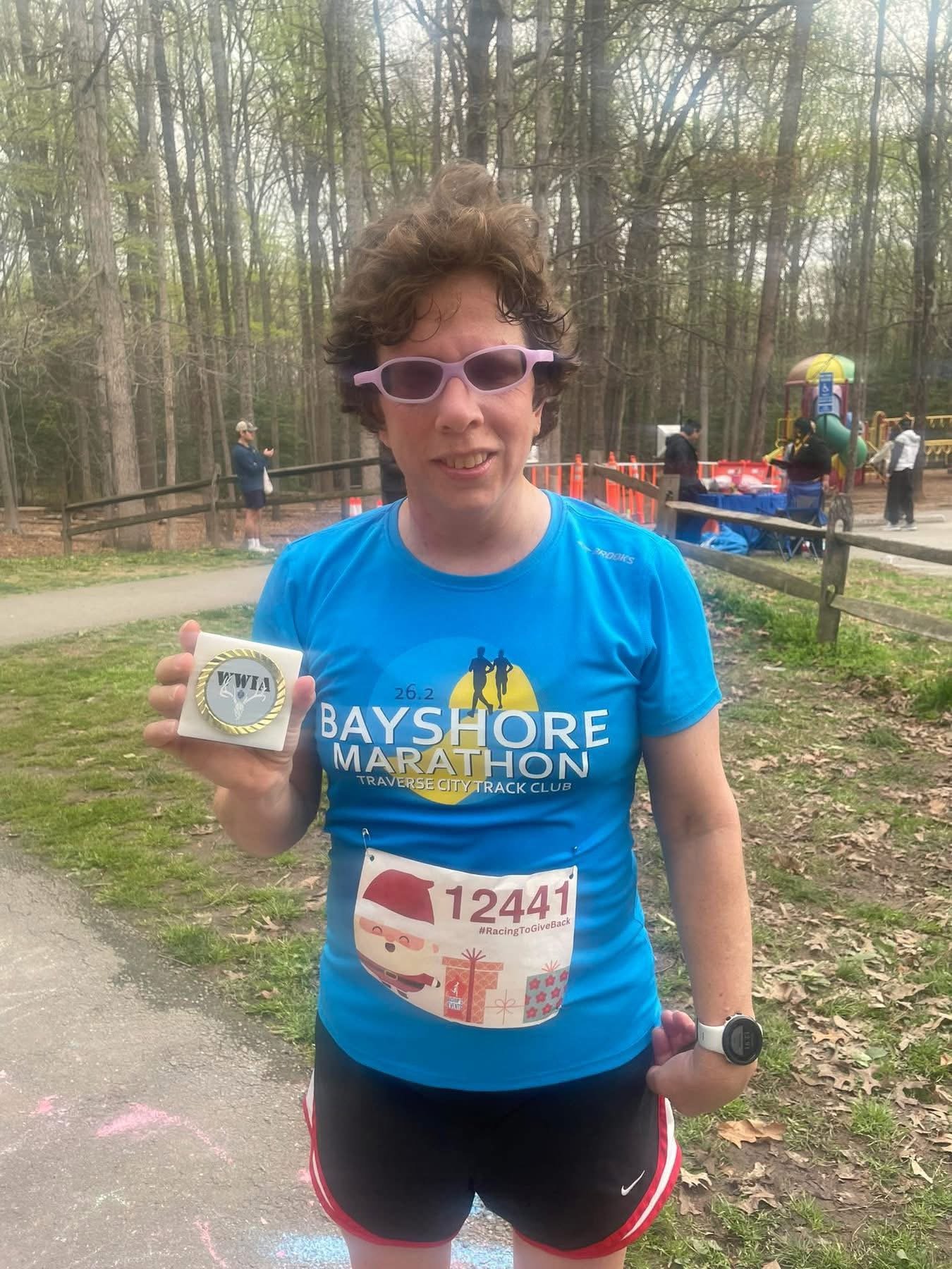 A woman in a blue Bayshore Marathon shirt holding a medal and standing outdoors at a park with trees and a playground in the background.