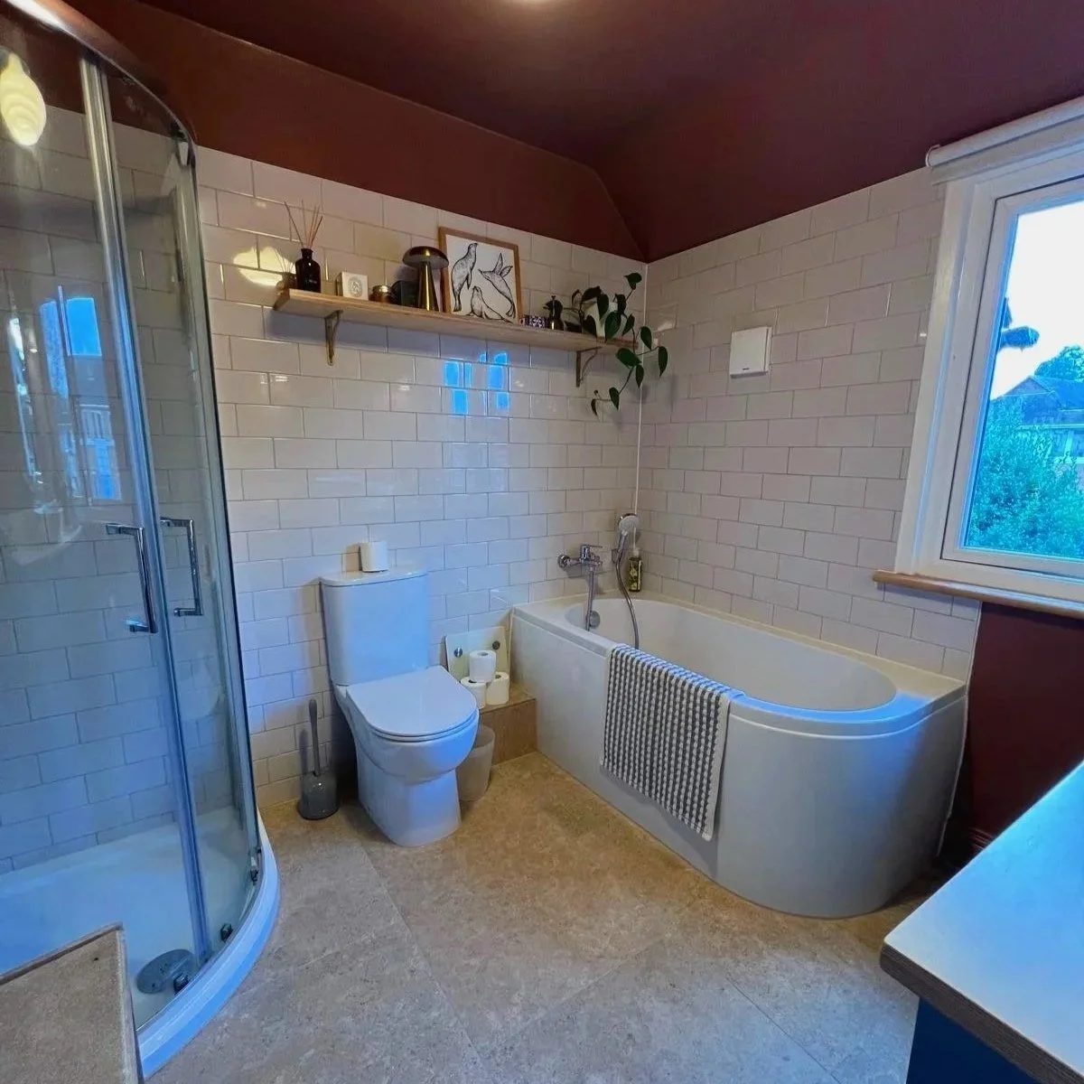 Bathroom with a corner shower, toilet with extra toilet paper, bathtub with a checkered towel, window, and a wall shelf decorated with plants, artwork, and decorative objects.