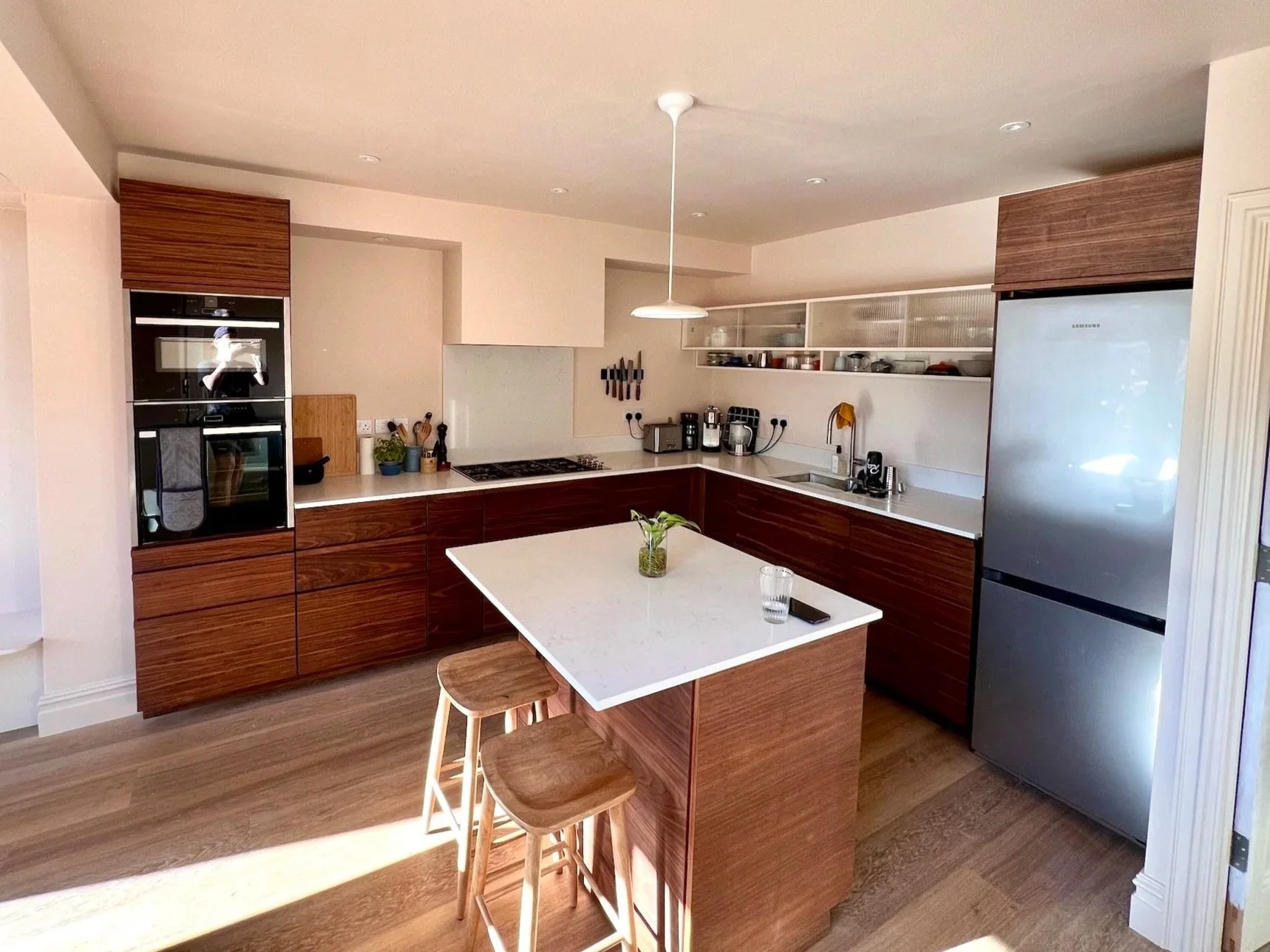 Modern kitchen with wood cabinets, a white island with wooden stools, and stainless steel appliances, including a refrigerator and oven.