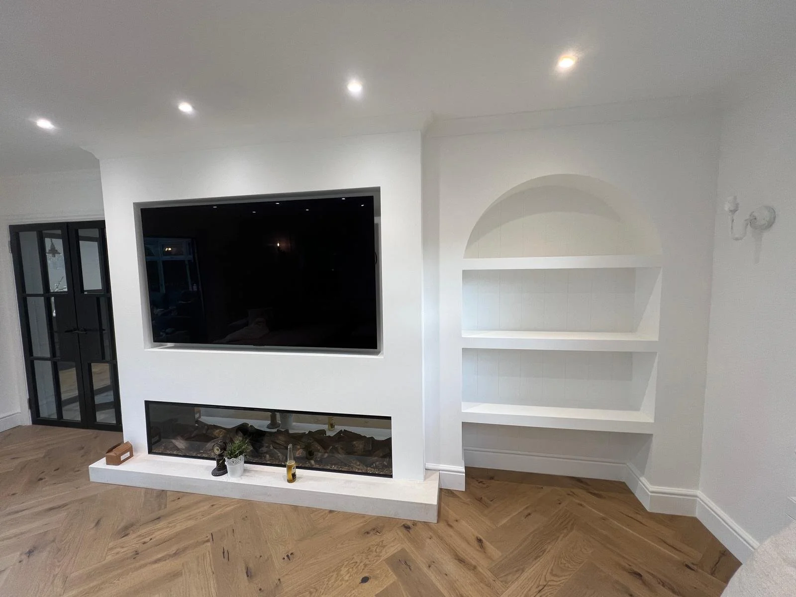 Living room with a wall-mounted flat-screen TV above a built-in fireplace, next to white built-in shelves with an arched top. The room has hardwood flooring and white walls, with black-framed glass doors on the left and ceiling lights.