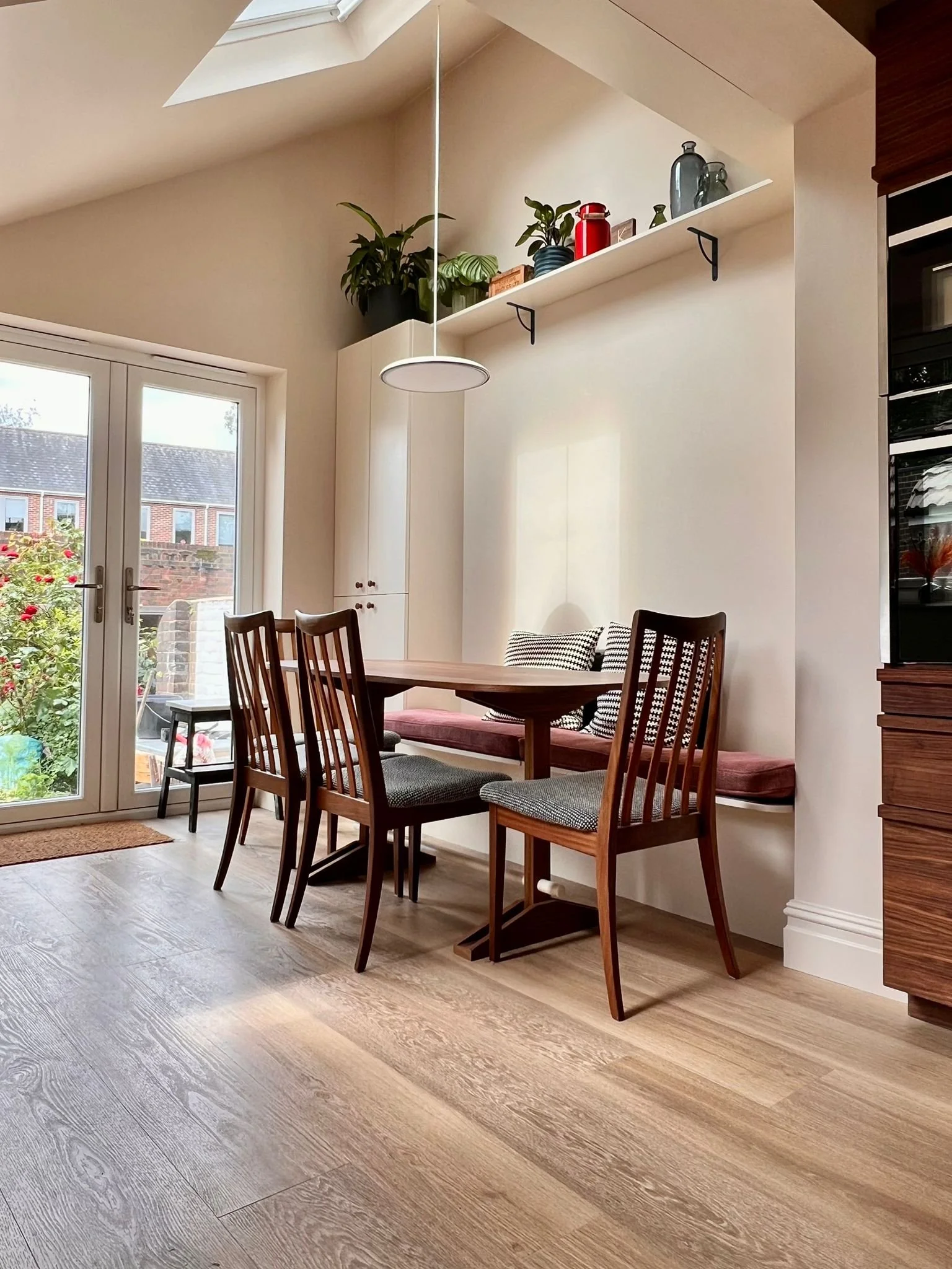 A cozy dining area with a wooden table surrounded by four wooden chairs, with two additional chairs along the wall, near a glass door leading to a garden. There are houseplants and decorative vases on a wall shelf above the table, and a skylight prov
