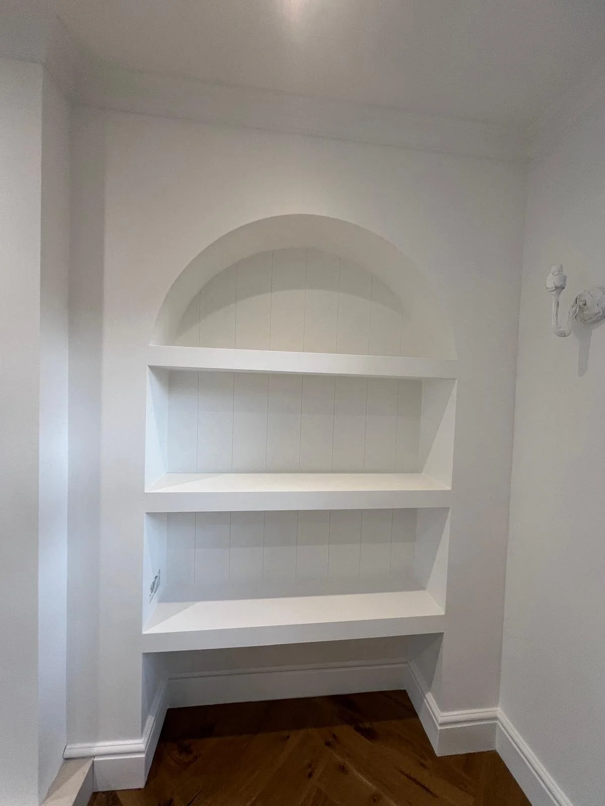 White built-in wall shelving with three open compartments, framed by an arched niche, located in a room with hardwood flooring and white walls.