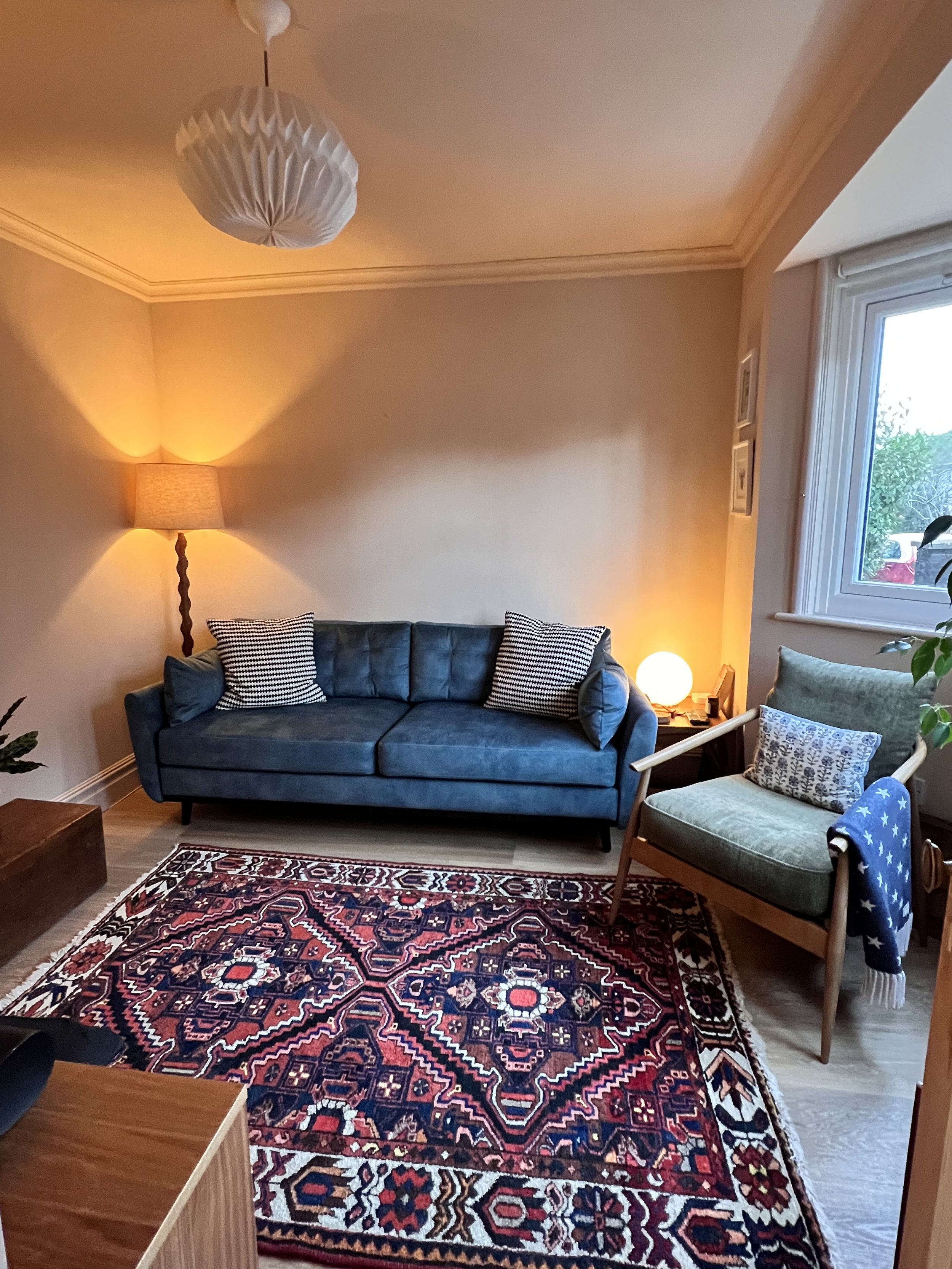 A cozy living room with a dark blue sofa, two striped pillows, a wooden side table with a spherical lamp, a green armchair with a decorative cushion and a navy blanket, a patterned rug, a standing lamp with a twisted pole, and a window with a view of