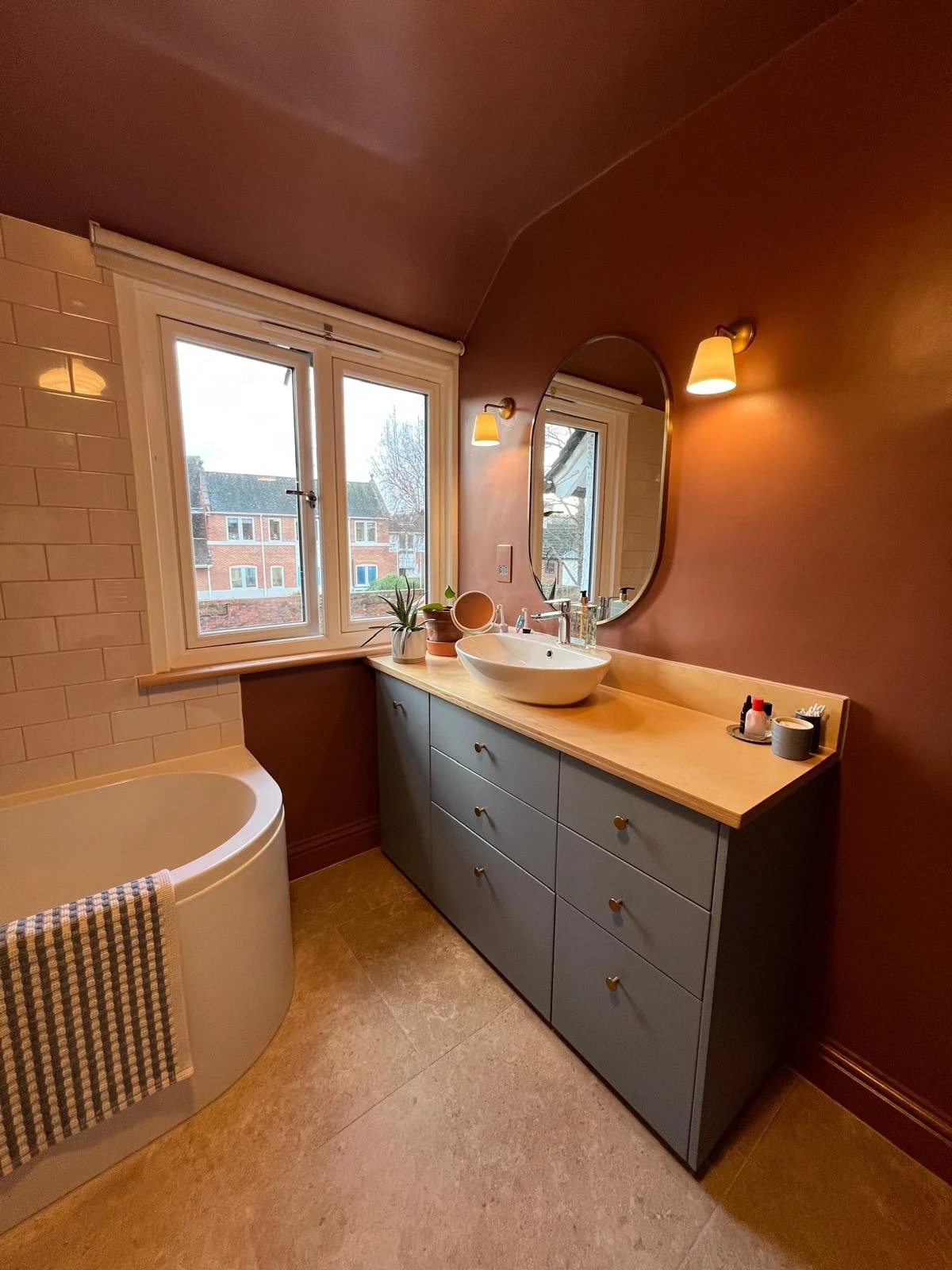 Modern bathroom with brown painted wall, large window, grey vanity with wooden countertop, white vessel sink, oval mirror, wall-mounted lights, bathtub, small plant, and bathroom toiletries.