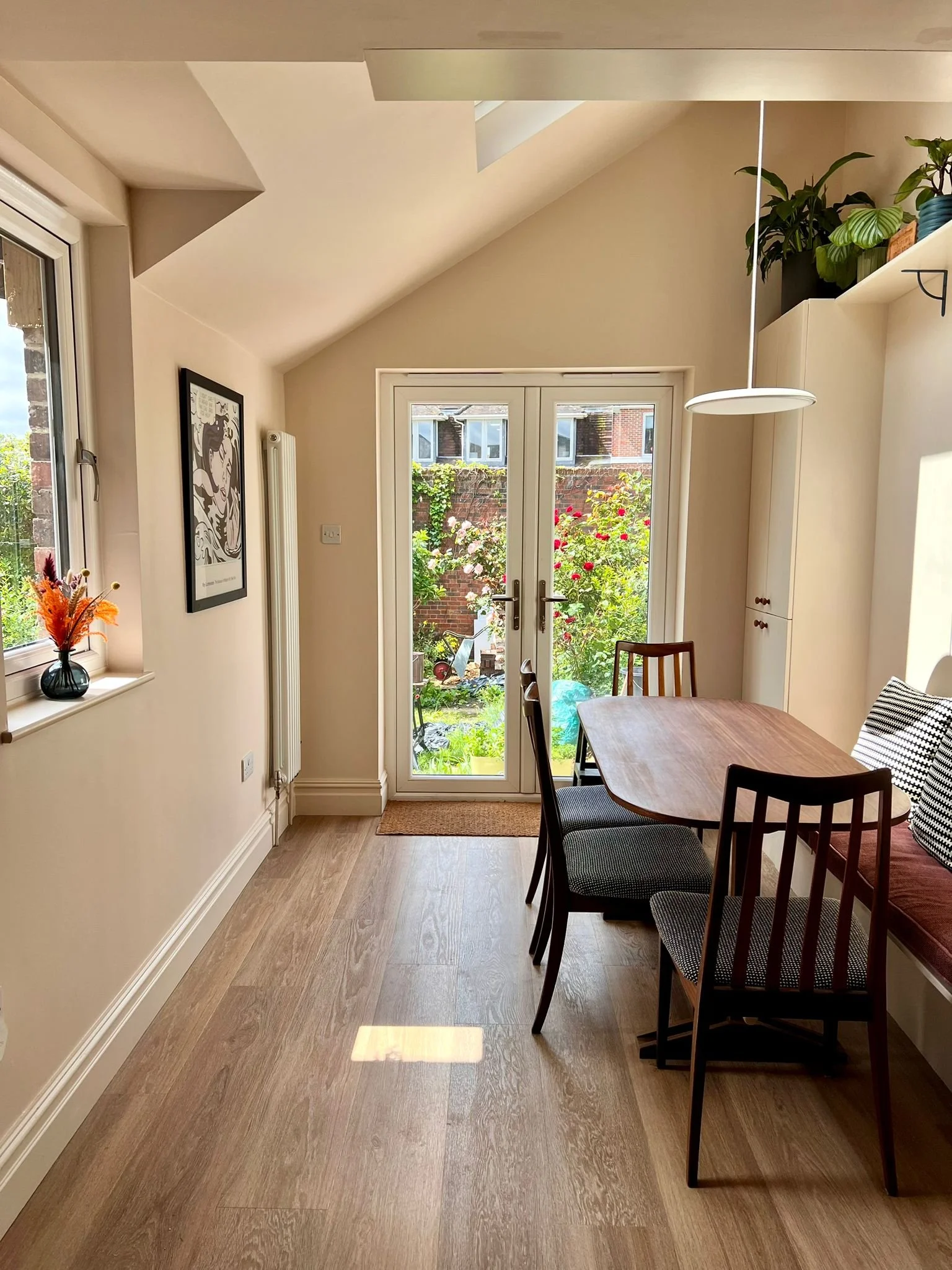 Dining area with a wooden table, chairs, a bench with pillows, a window sill with a vase of flowers, a door leading to a garden, and potted plants on a shelf.