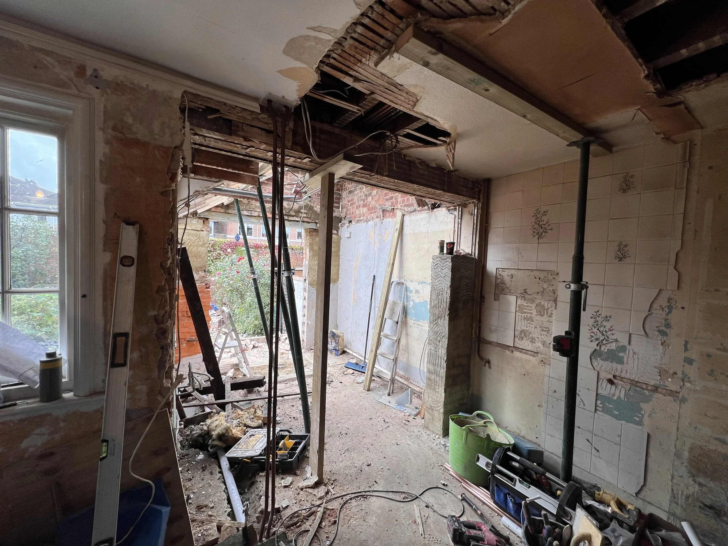 Room under renovation with exposed walls and ceiling, construction tools, and scaffolding. Outside view through an opening shows a garden with flowers.