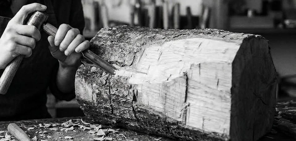 Person carving into a large tree trunk with a chisel and hammer in a woodworking workshop.