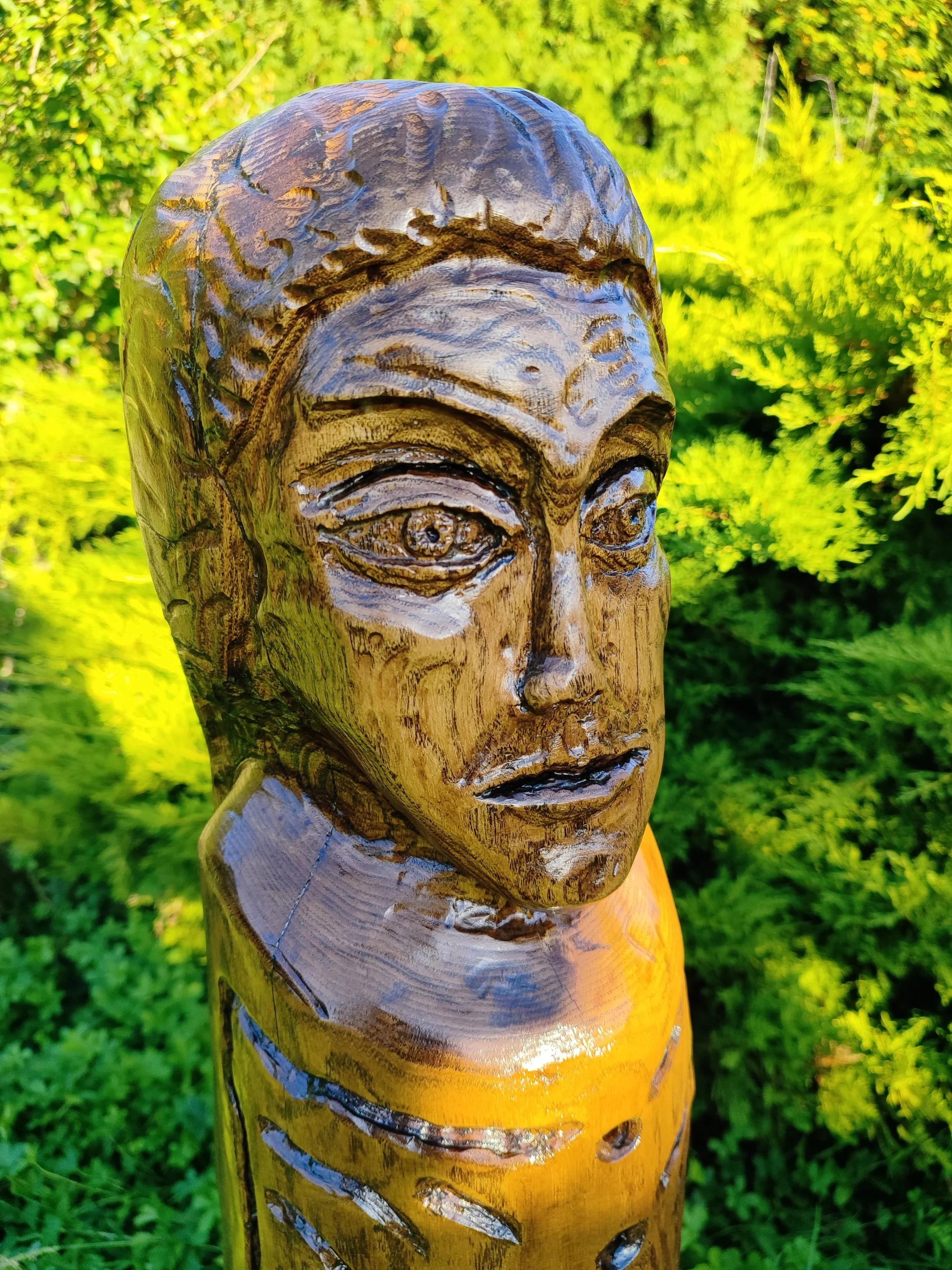 Close-up of a carved wooden sculpture of a human face on a wooden post, set outdoors with green foliage in the background.