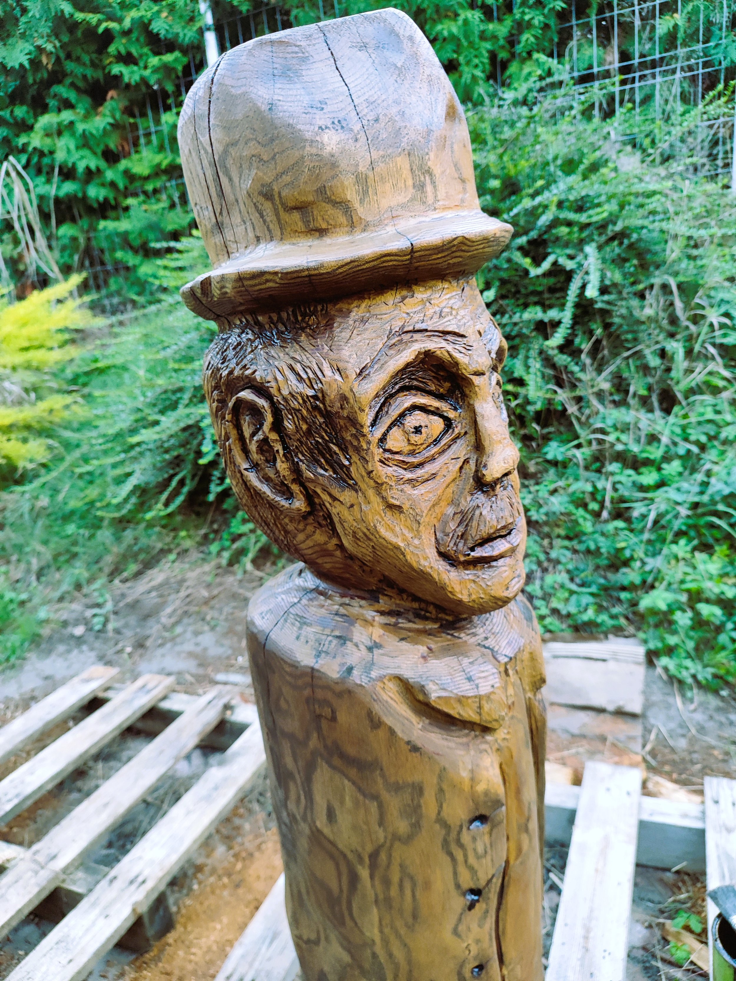 A wooden sculpture of a human face with an expressive look, wearing a hat, surrounded by green bushes and trees, with a wooden handrail nearby.