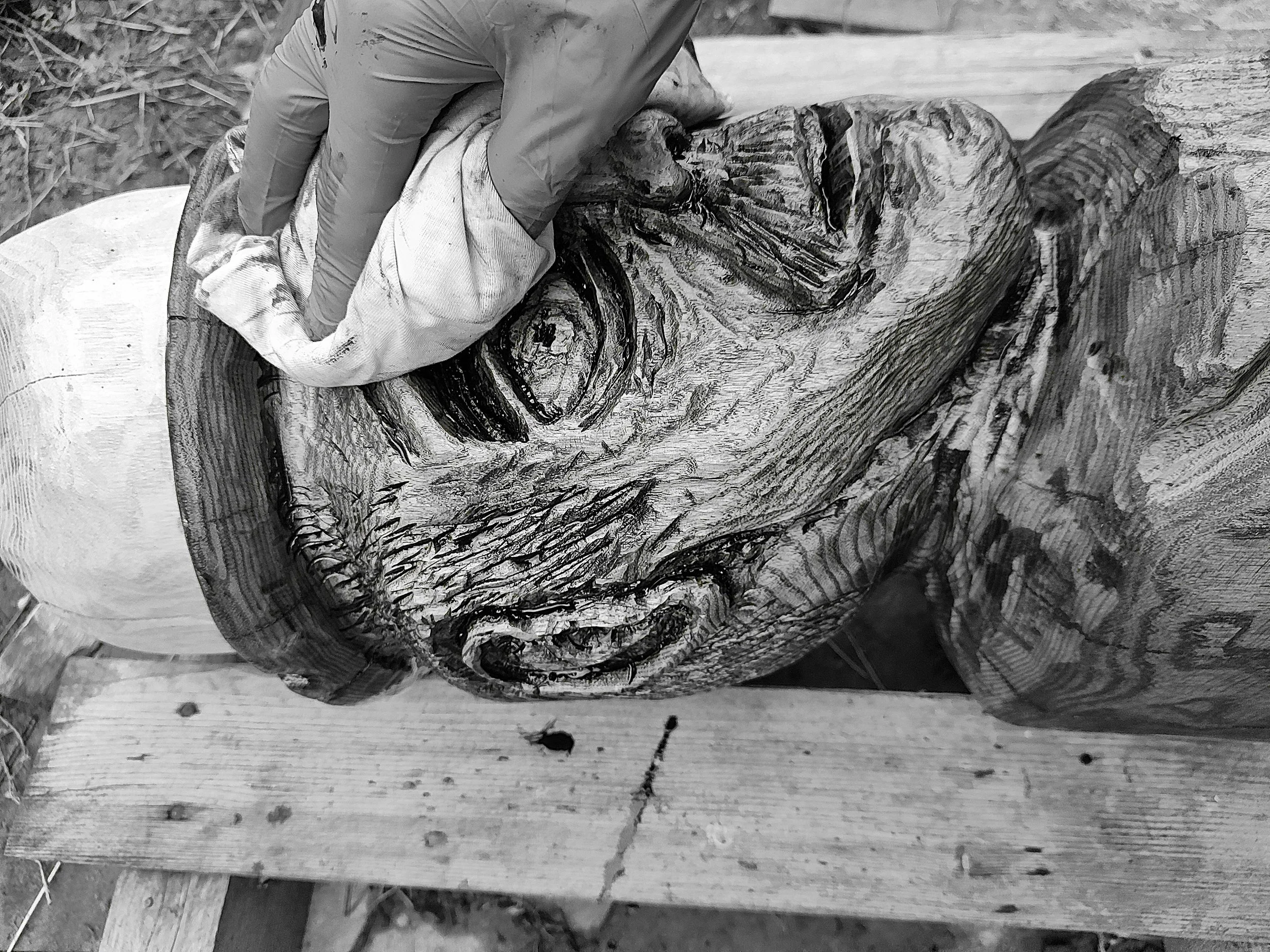 A person wearing gloves carving or shaping a large piece of wood or log, with visible wood grain and texture, on a wooden surface outdoors.