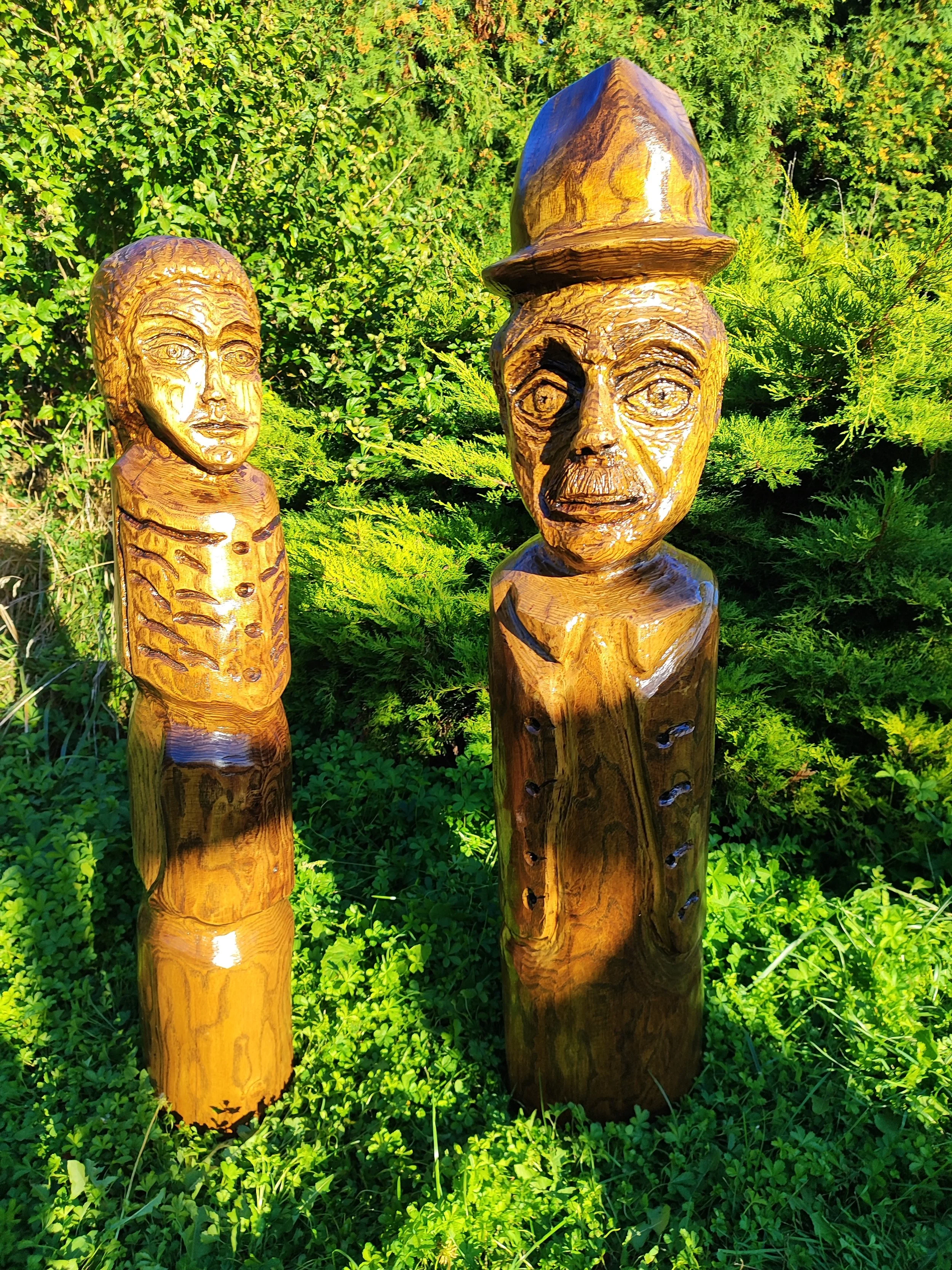 Wooden sculptures of a boy with a hat and a girl with a ponytail, set outdoors with green foliage in the background.