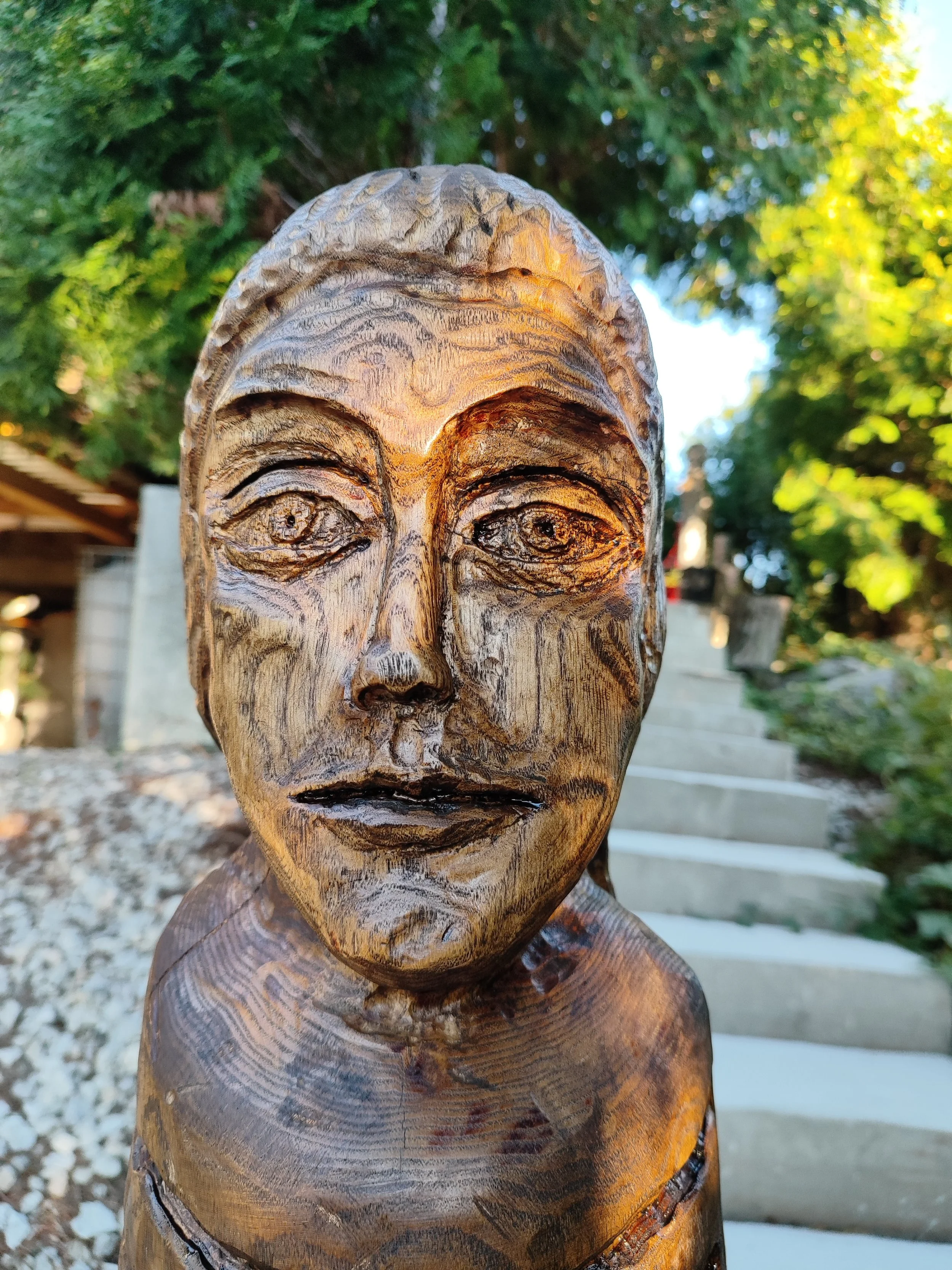 Close-up of a wooden sculpture of a human face with detailed features, set outdoors with trees and stairs in the background.