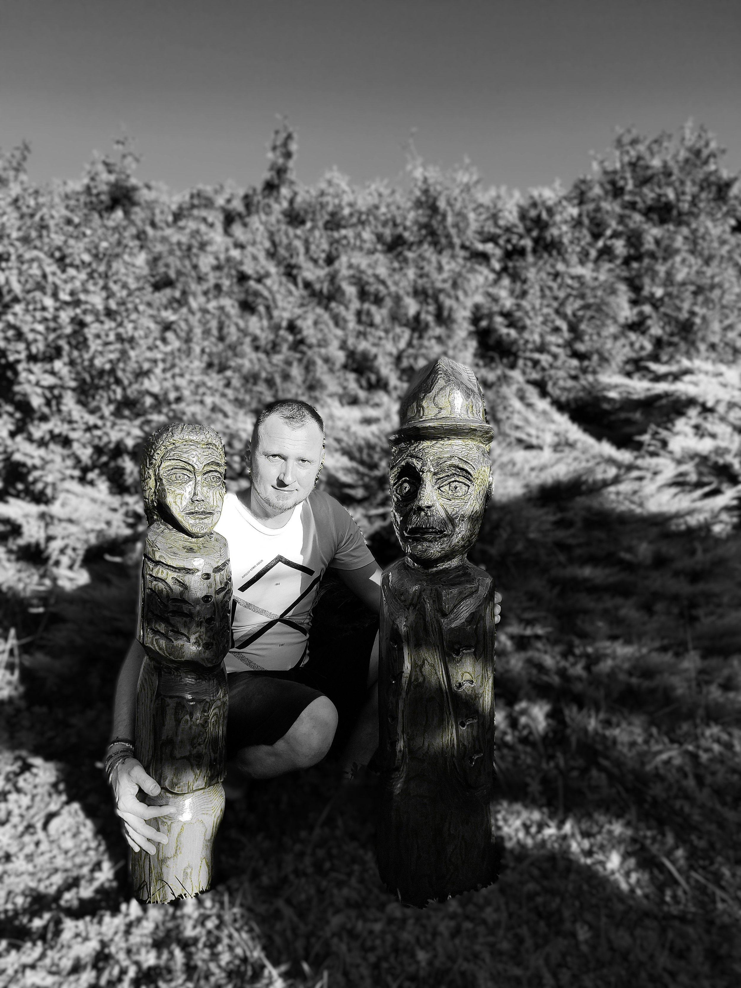 A man squatting outdoors, holding two wooden sculptures of faces, with a background of trees and grass.