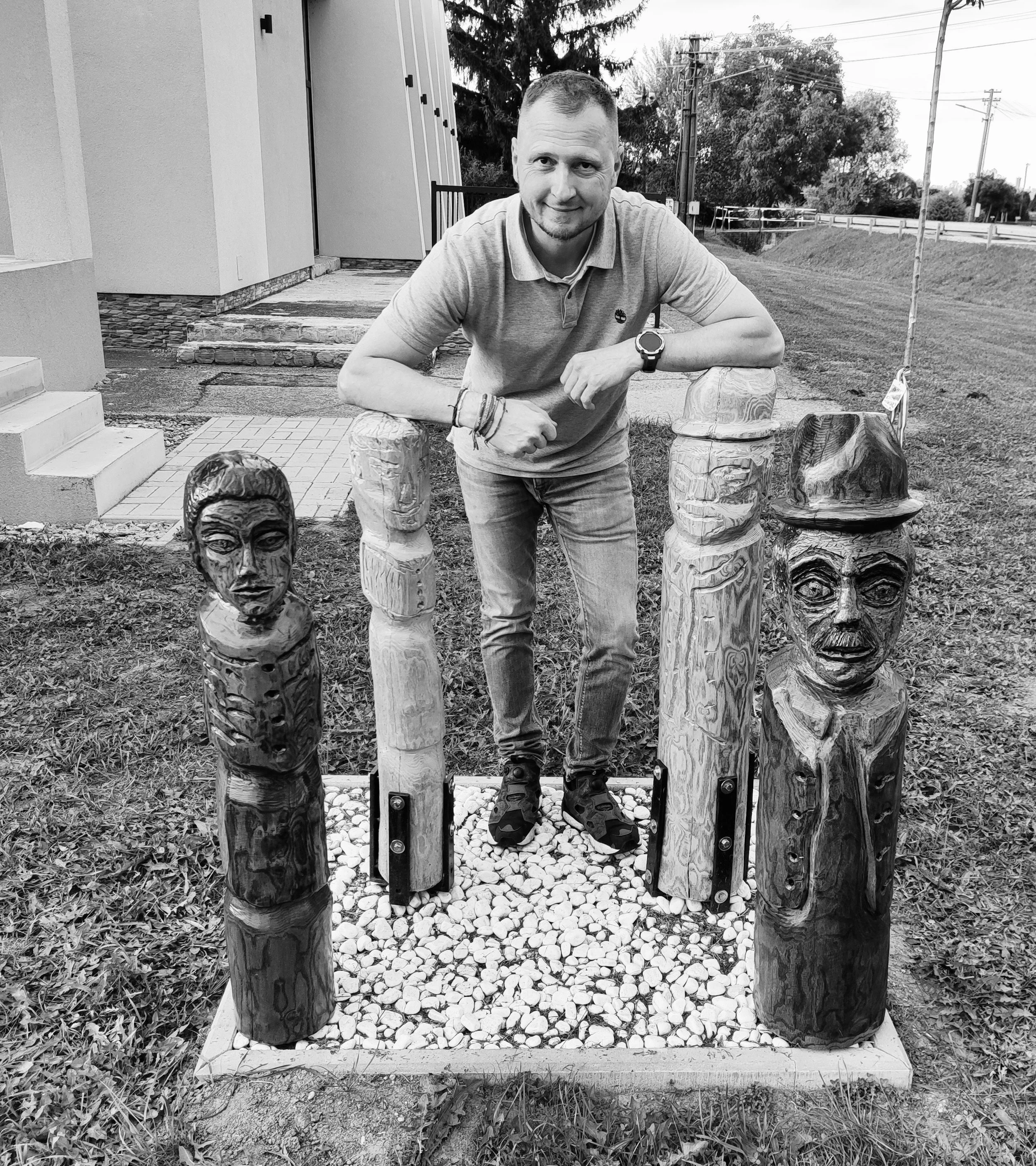 A man leaning on wooden carved totem poles outside near a house, with trees and utility poles in the background. The image is in black and white.