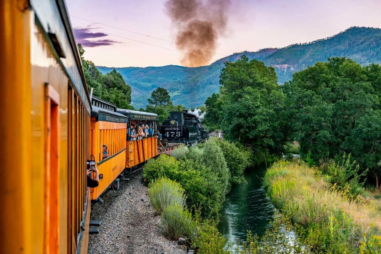 A vintage train traveling through a lush green landscape with a river on the side, mountains in the background, and a plume of dark smoke rising from the engine, during sunset.