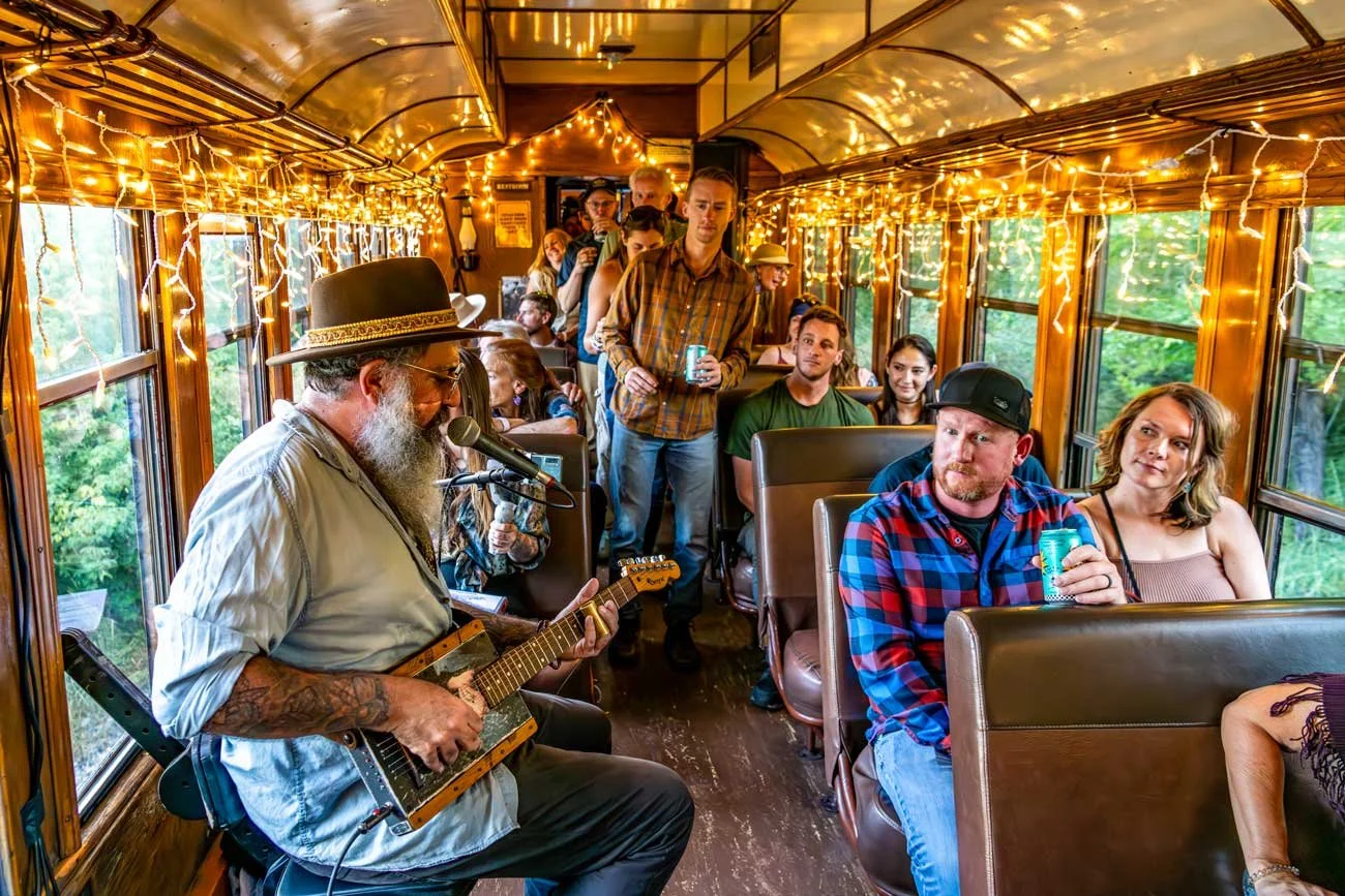 A man with a gray beard wearing a large hat and glasses playing a guitar and singing into a microphone inside a decorated vintage train car with string lights, while seated passengers listen, some holding drinks.