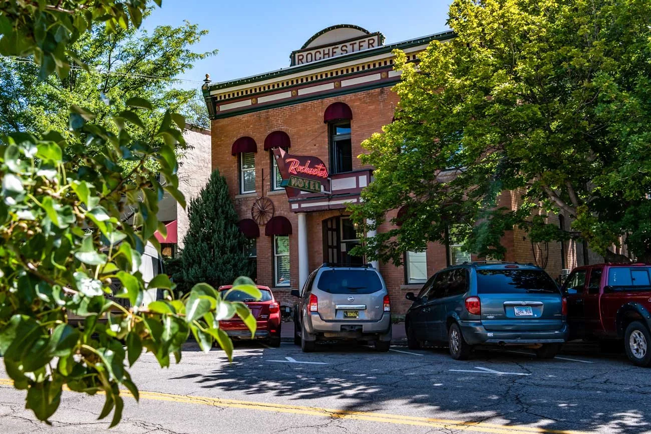 A historic brick building with a neon sign that reads 'Rochester' and a smaller sign that says 'Rochester Motel.' There are several parked cars in front of the building, surrounded by lush green trees, on a sunny day.