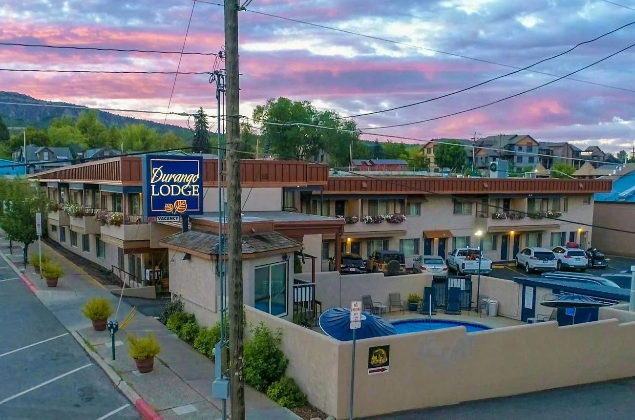 A two-story motel named Durango Lodge with a sign showing vacancies, parking lot with several cars, small pool area with lounge chairs, and surrounding residential houses under a colorful sunset sky.