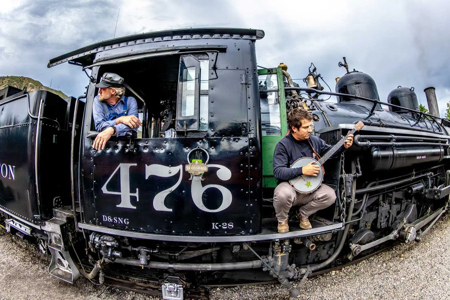 Two men on a vintage black steam locomotive, one sitting with a banjo and the other looking away, with a scenic outdoor background.
