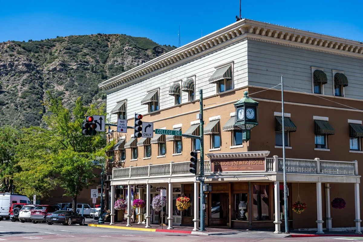 Corner view of the General Palmer Hotel on Collage Drive, with street signs, hanging flower baskets, a clock, and a mountain in the background.
