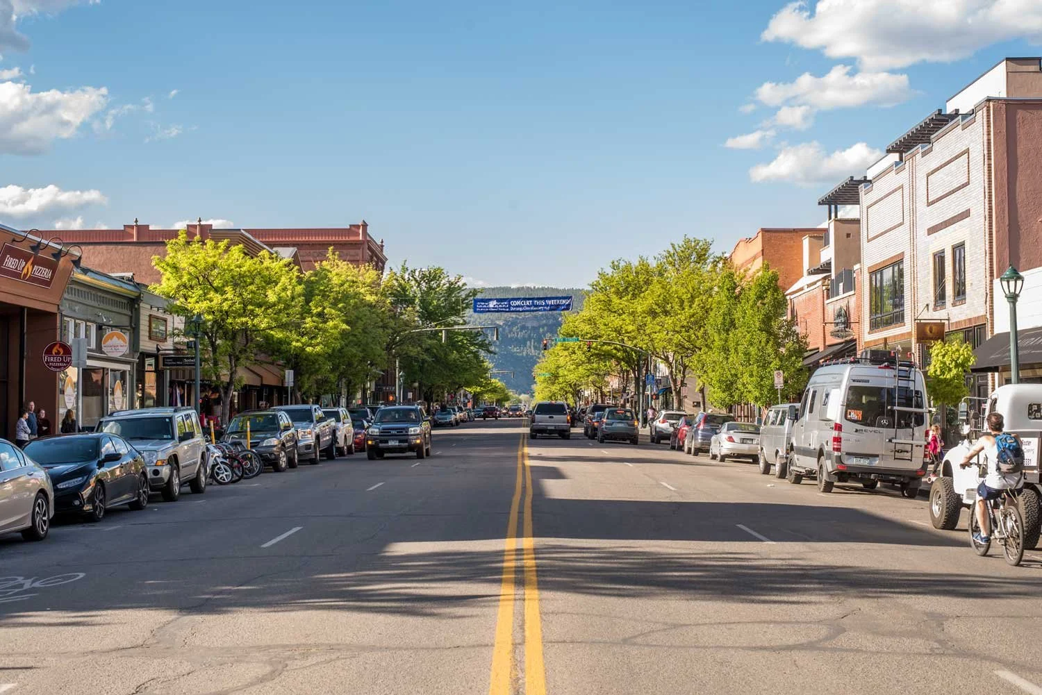 A city street with cars parked along both sides, trees lining the sidewalks, and a mountain in the background under a blue sky with clouds.