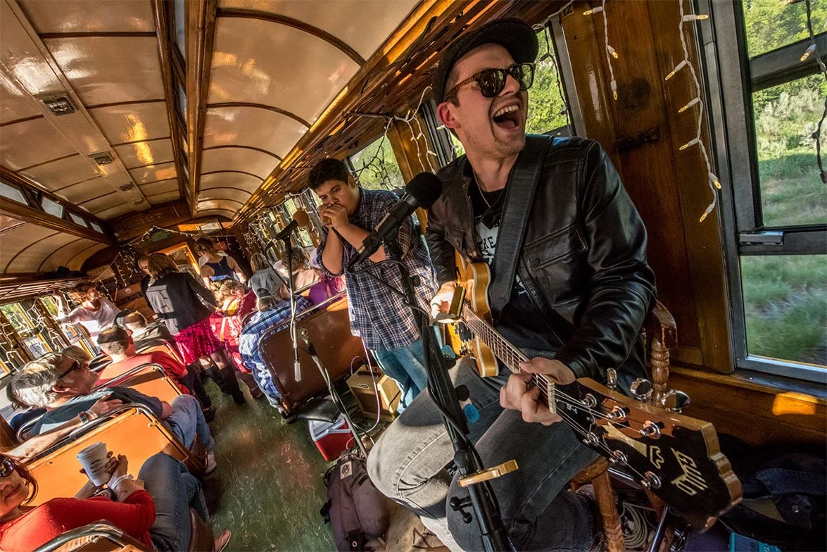 A man wearing sunglasses and a leather jacket plays an acoustic guitar and sings into a microphone on a train with wooden interior. Several passengers are sitting and standing, some wearing hats, enjoying the music. Sunlight streams through windows, illuminating the scene.