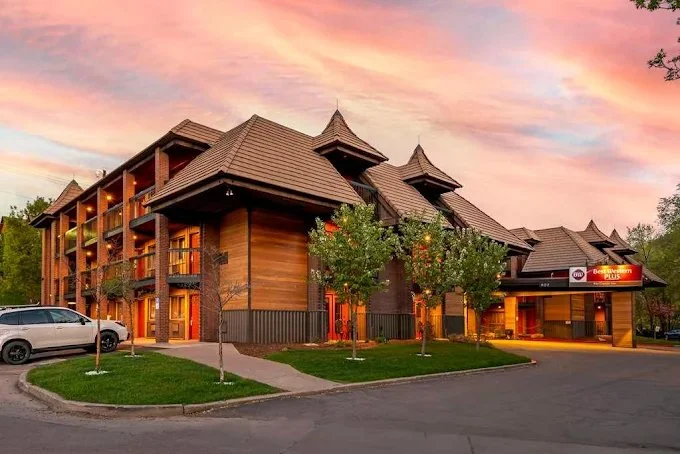 A modern hotel with wooden accents and multiple balconies, surrounded by young trees, under a pink, orange, and purple sunset sky.