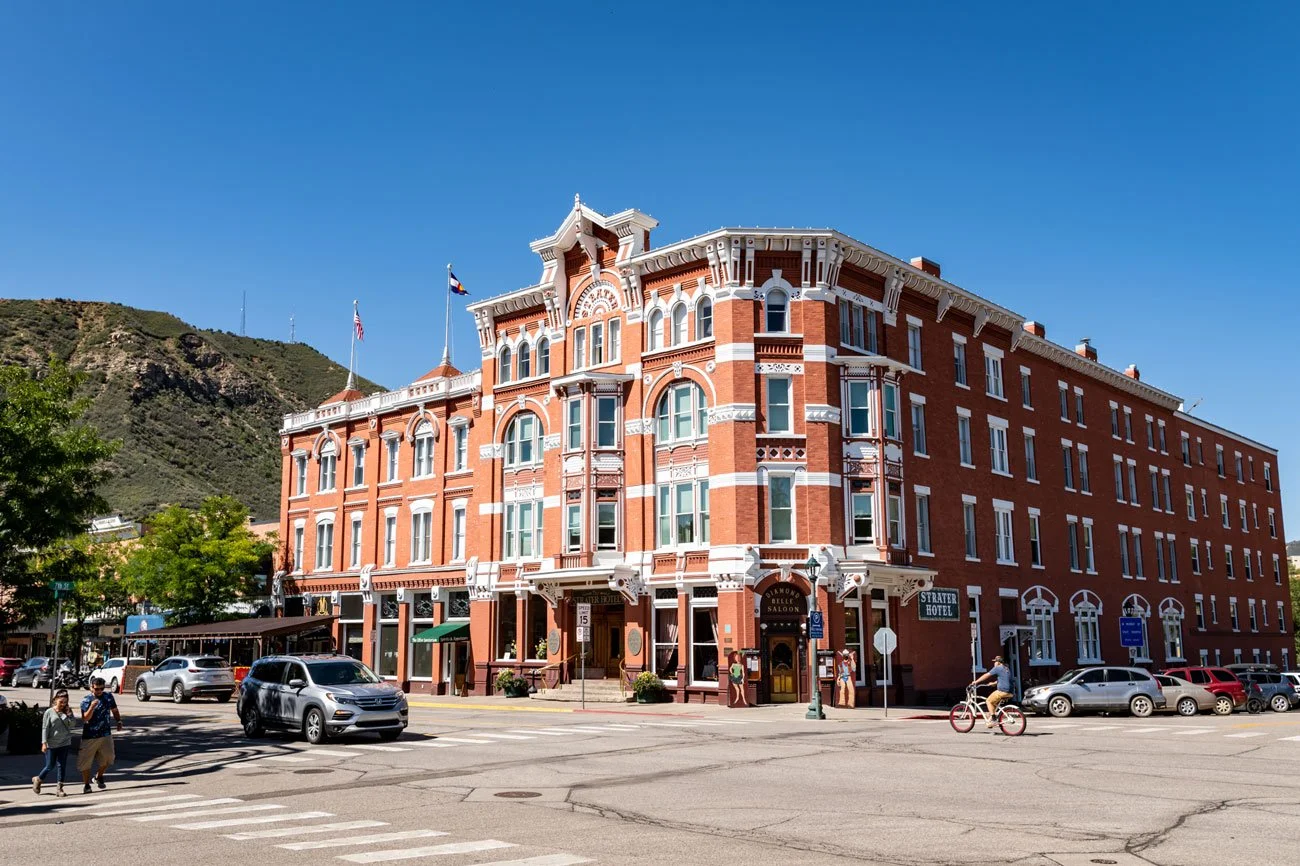 Historic red brick building with white architectural details, located on a street corner under a clear blue sky, with cars parked and people walking or biking nearby.