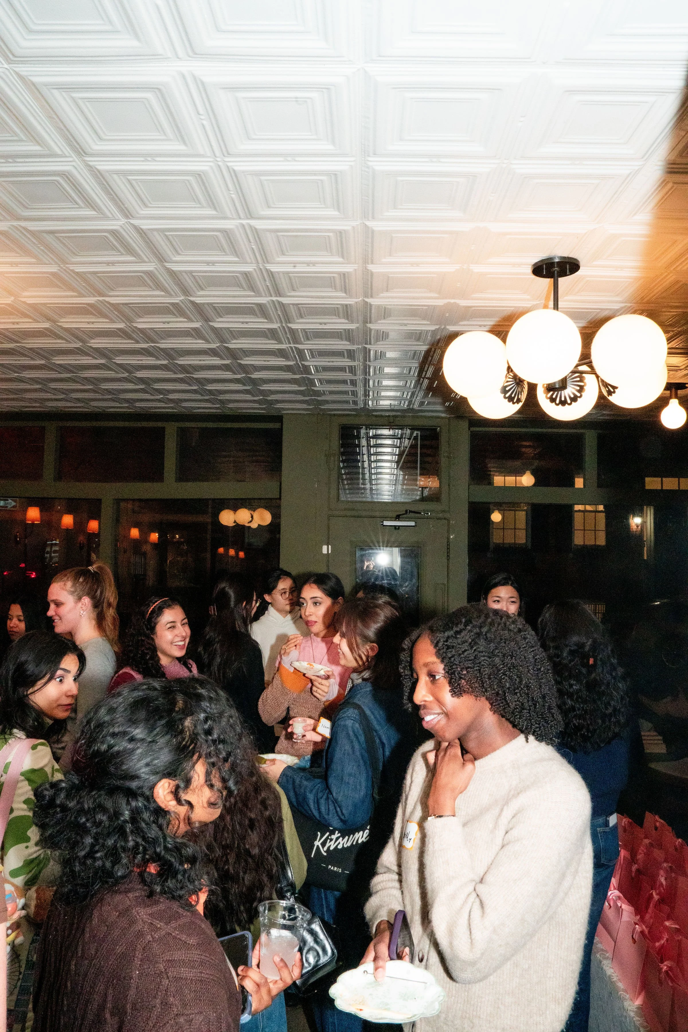 Group of women socializing in a warmly lit indoor space with decorative ceiling tiles and round ceiling lights. Girls Gone Hiking Gutsy Galentine's hosted at Bedford Studio in the West Village NYC.