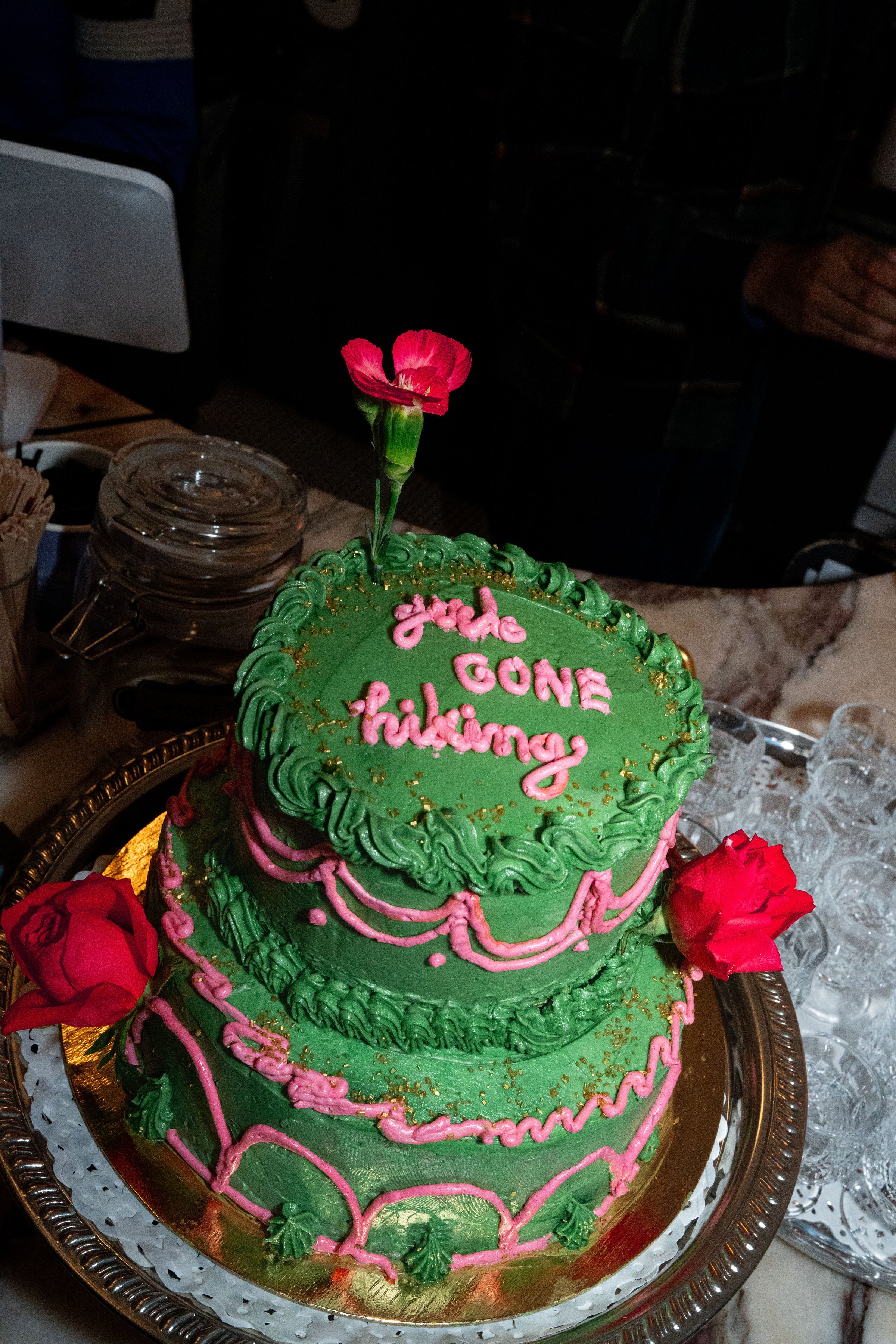 A three-tier green cake decorated with pink icing accents, red roses, and pink scripting that reads 'Girls Gone Hiking'