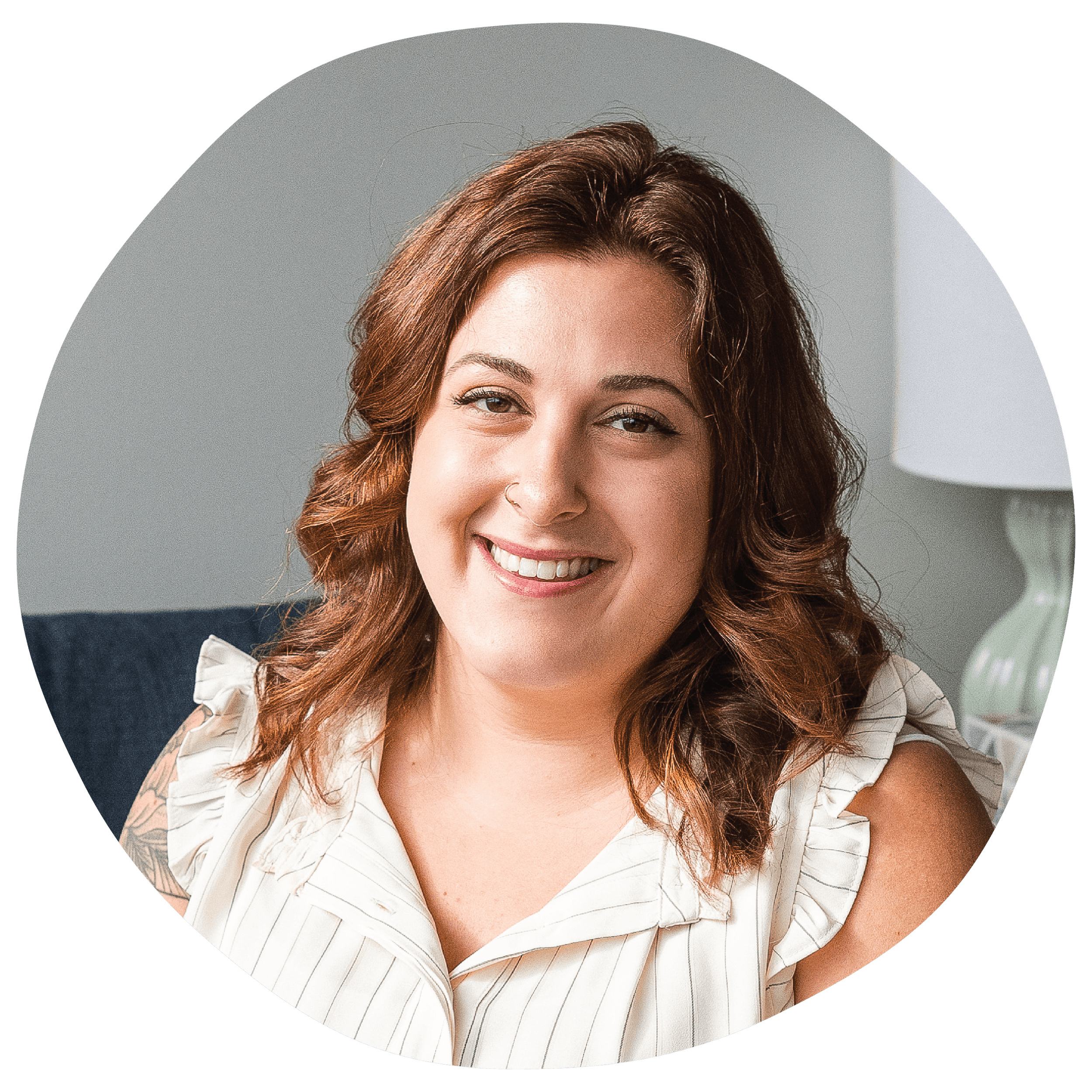Smiling woman with shoulder-length wavy brown hair, wearing a white sleeveless top, sitting indoors near a lamp.