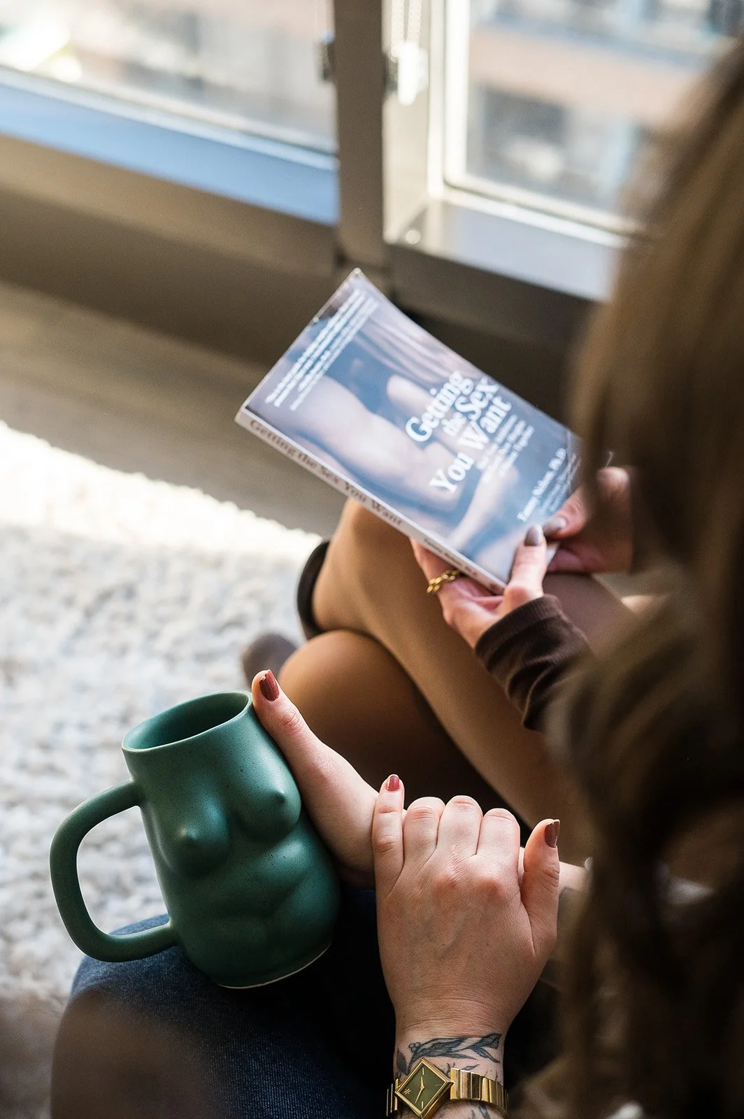 A person sitting by a window reading a book called "Getting The Sex You Want", holding a large green mug with a torso design, and wearing a gold watch and ring.