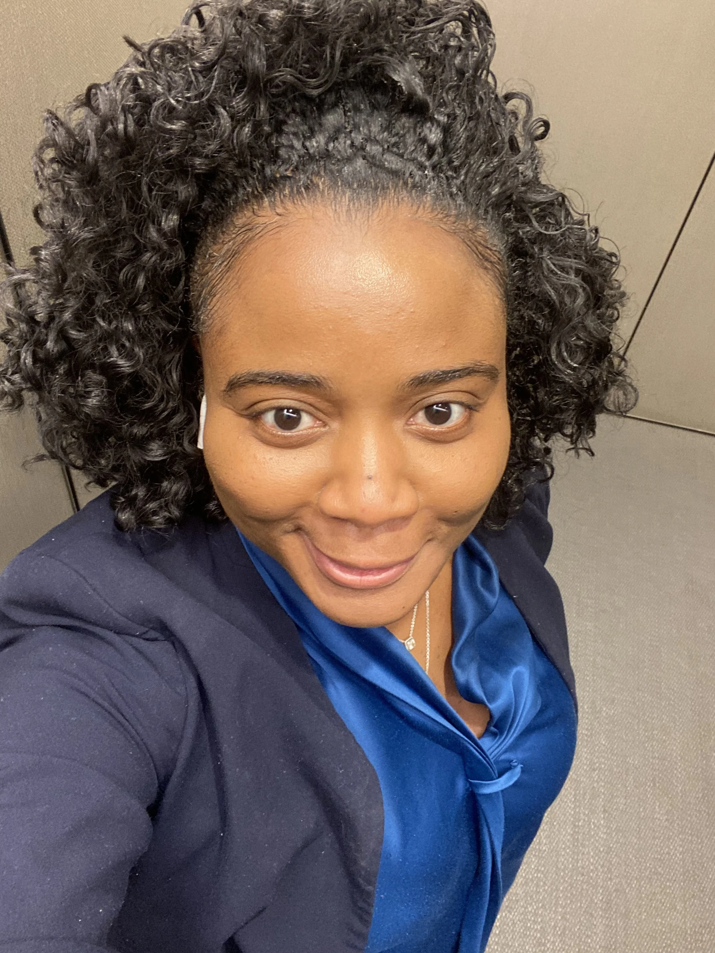 A woman taking a selfie from above, smiling, with curly hair, wearing a blue satin blouse and a dark blazer.