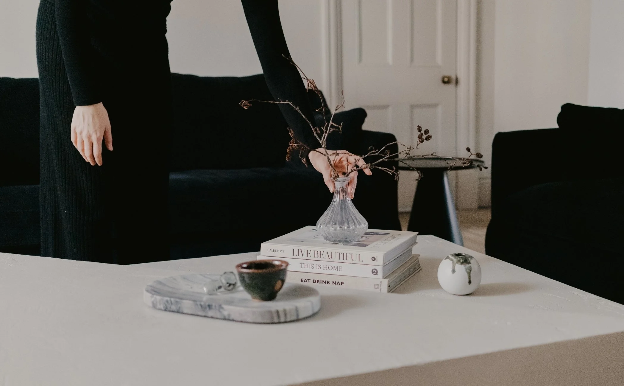 A person in black clothing adjusting a branch with small dried leaves in a glass vase on a stack of books on a white table. Items on the table include a small black bowl with a spoon, a white marble tray, and a white spherical object with gold accents. A black sofa and a white door are in the background.