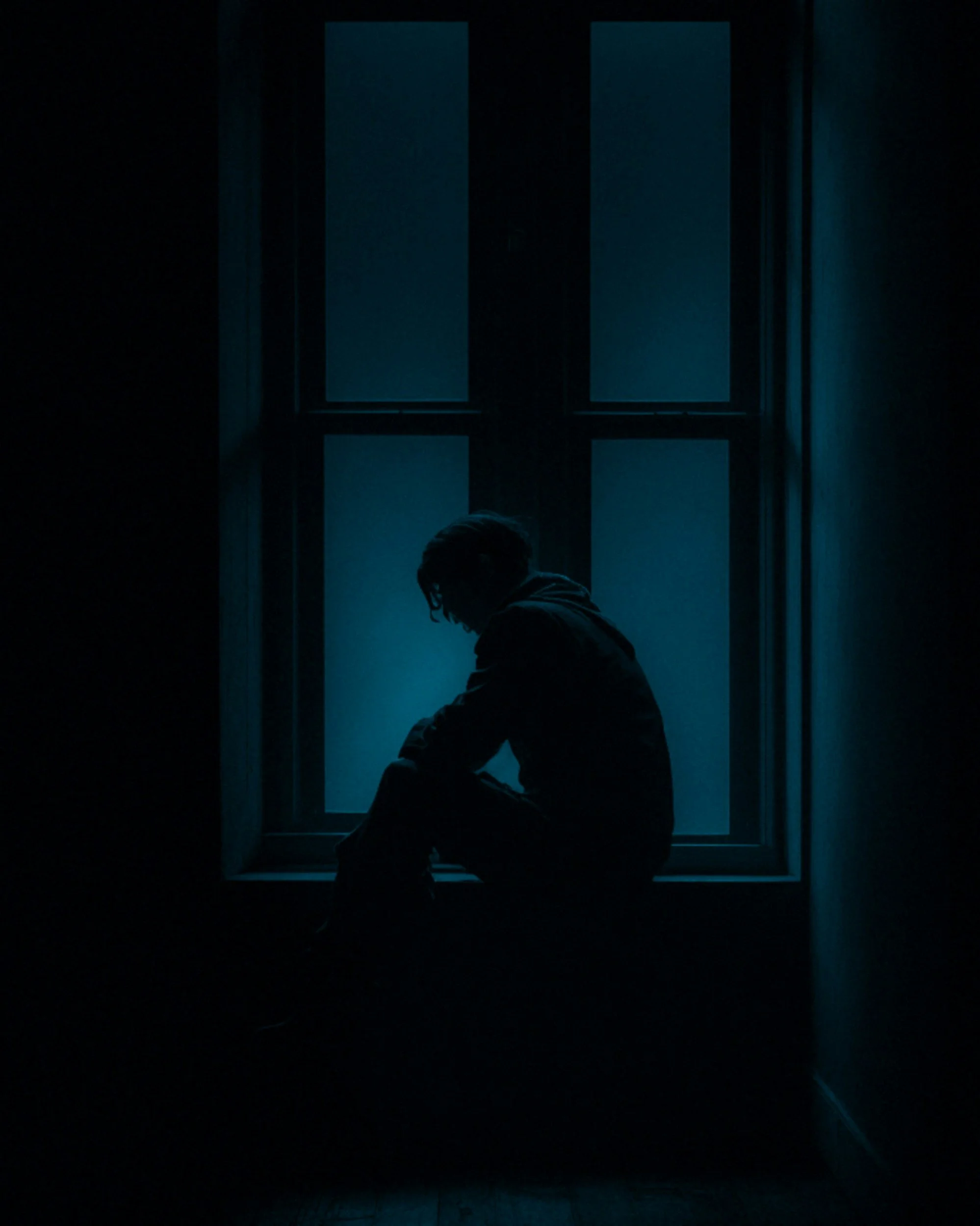 Silhouette of a person sitting on a windowsill at night, with blue light coming through the window.