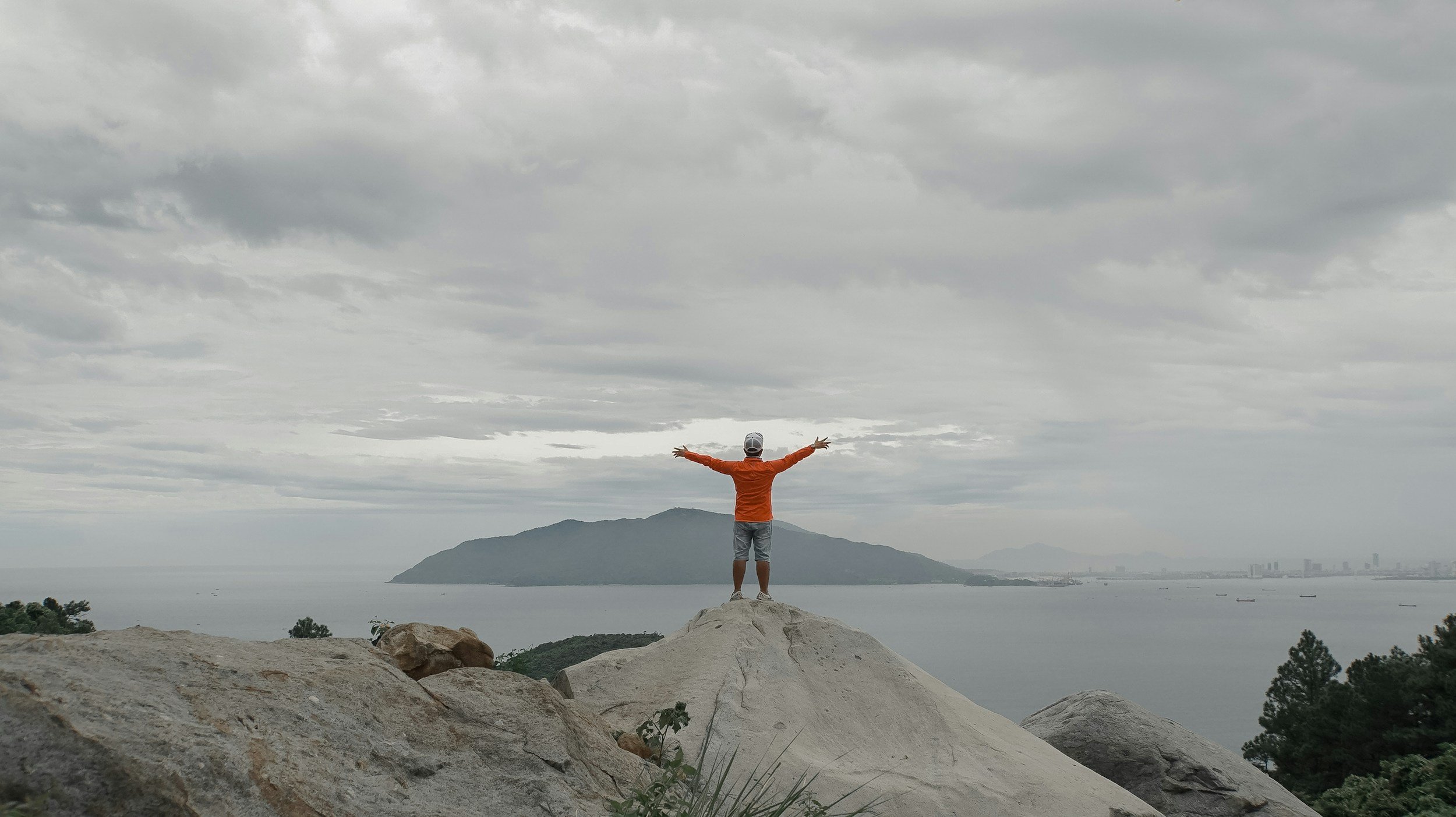 Person standing on a rocky hill with arms outstretched, overlooking a body of water and an island under a cloudy sky.
