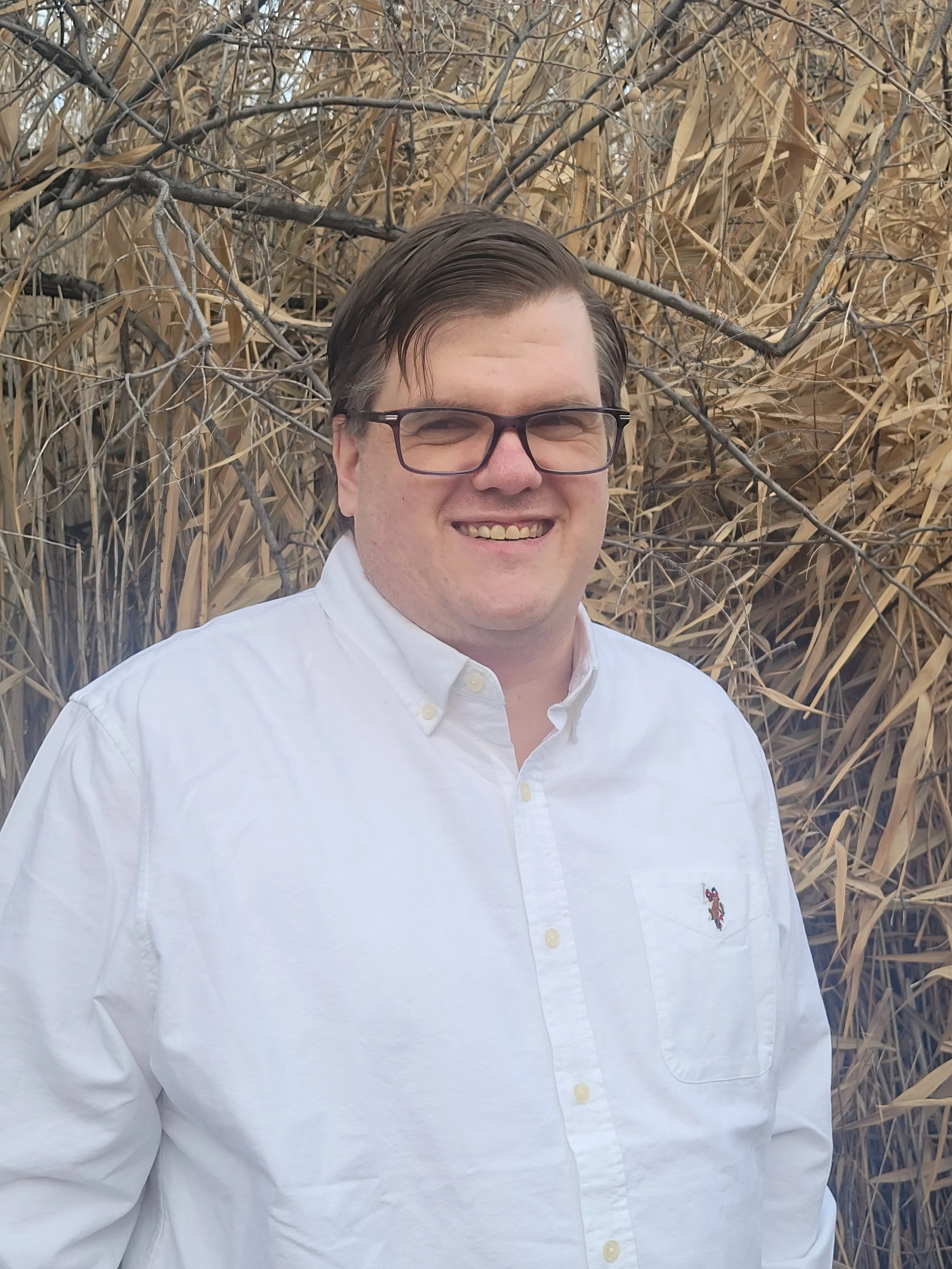 A man with glasses and dark hair smiling in front of tall, dry grasses or reeds.