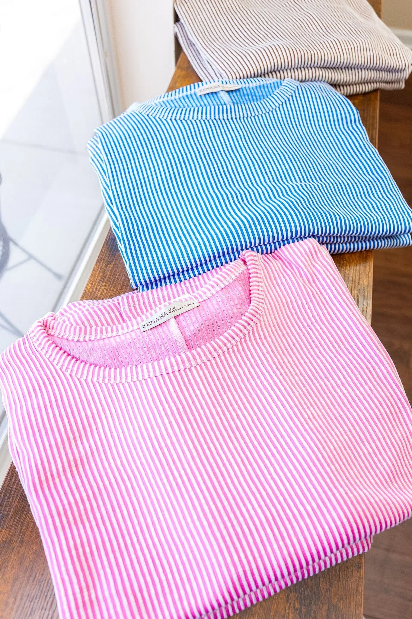 Three folded striped t-shirts in pink, blue, and black and white, placed on a wooden surface. These items can be found at The Soul Space Wellness Cafe + Lifestyle Shoppe in Panama City.