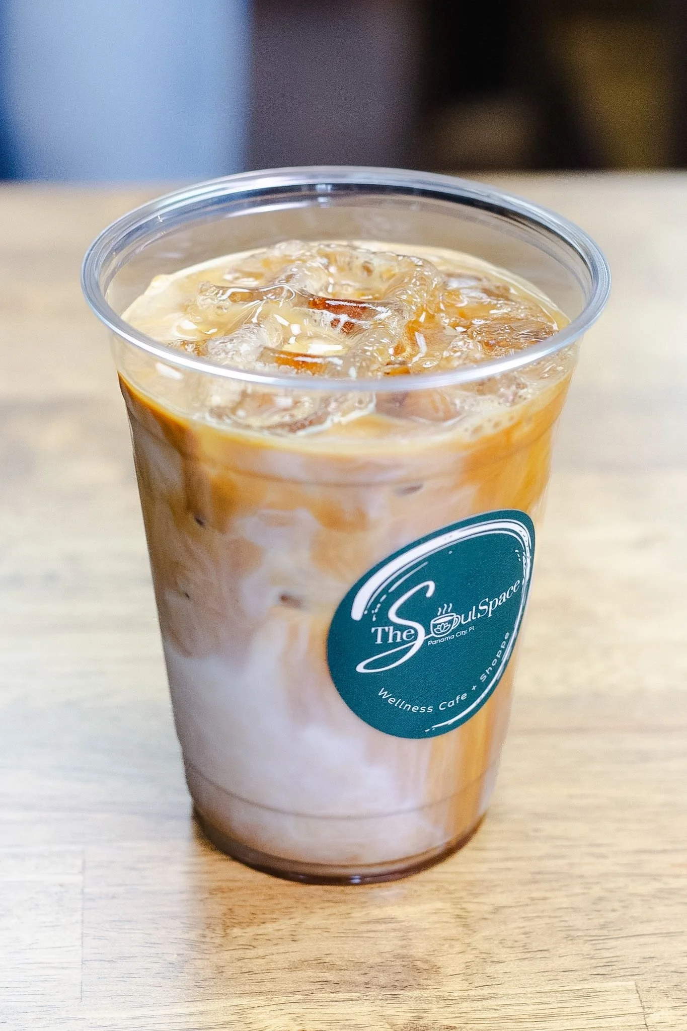 A clear plastic cup filled with iced coffee, showing swirls of milk, on a wooden surface. The cup has a teal circular logo that reads 'The Soul Space' and 'Wellness Cafe + Shop'.