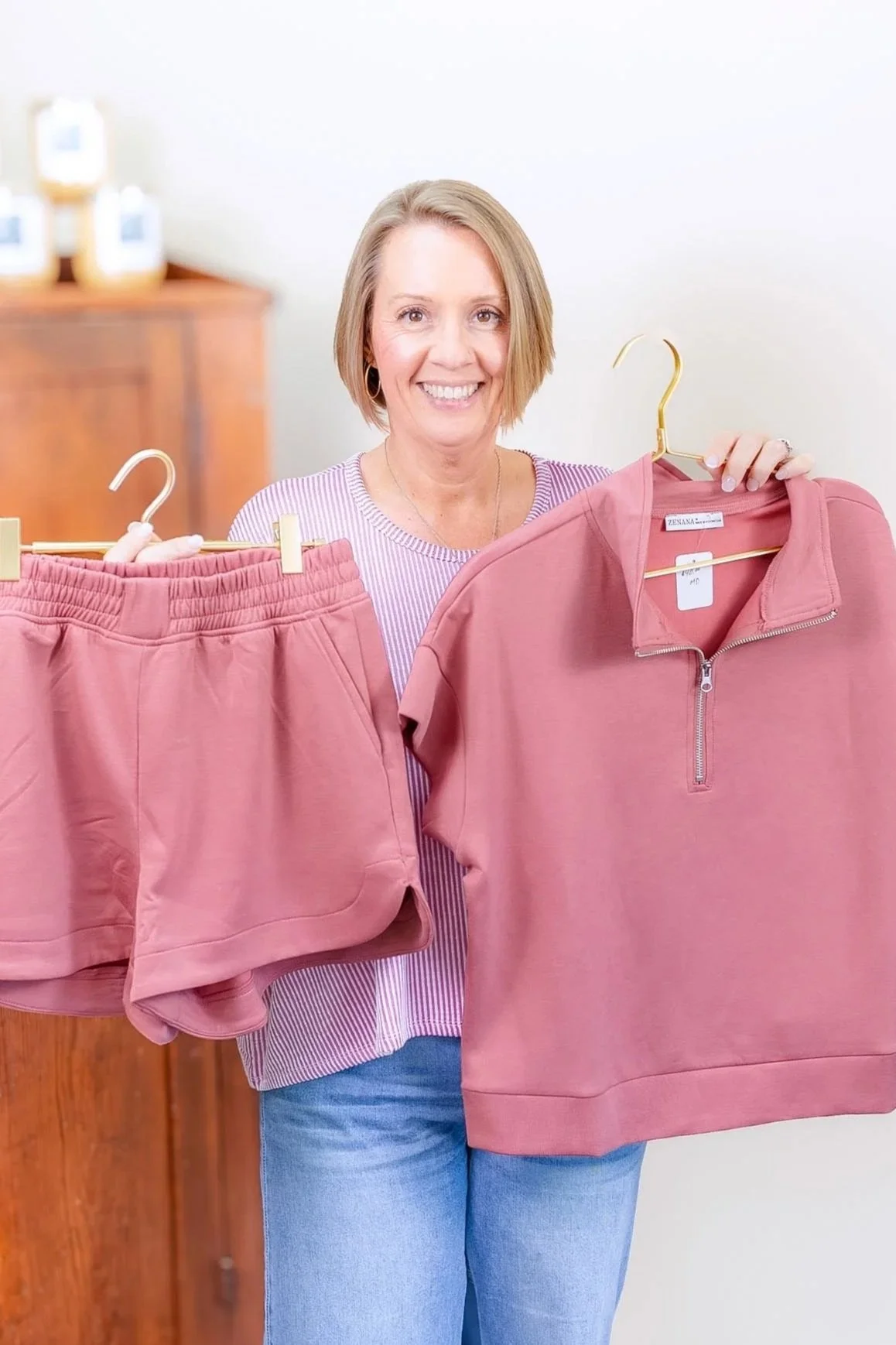 A woman smiling and holding pink loungewear/athleisure, including shorts and a sweatshirt, in a room with a wooden cabinet in the background. This boutique is located at The Soul Space in Panama City.