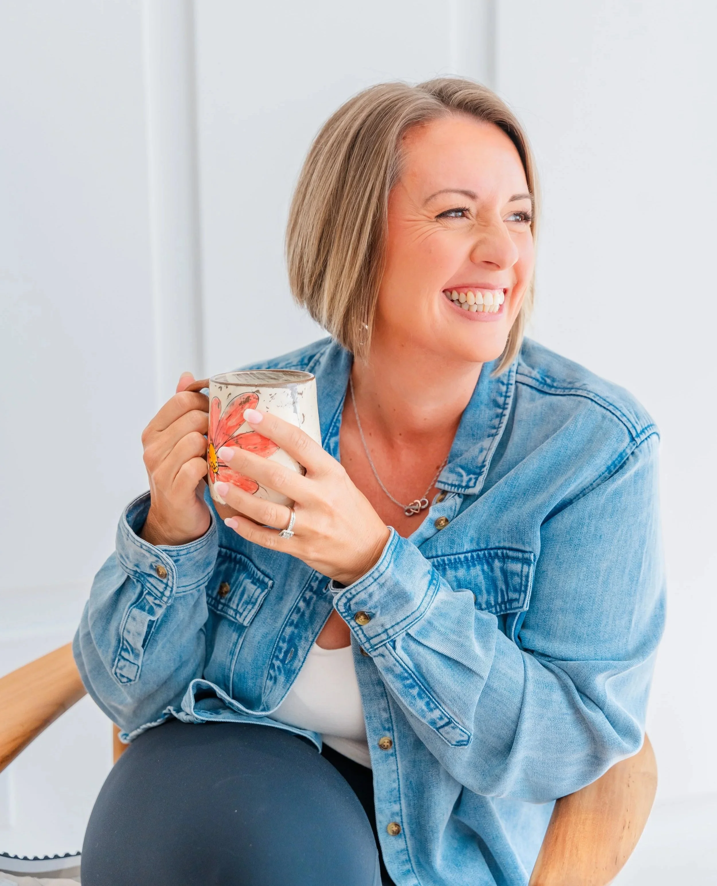 Woman sitting in a light, minimalist studio holding a floral mug and smiling, reflecting The Soul Space’s vision of creating a warm, welcoming place for community, wellness, and meaningful connection in Panama City, FL