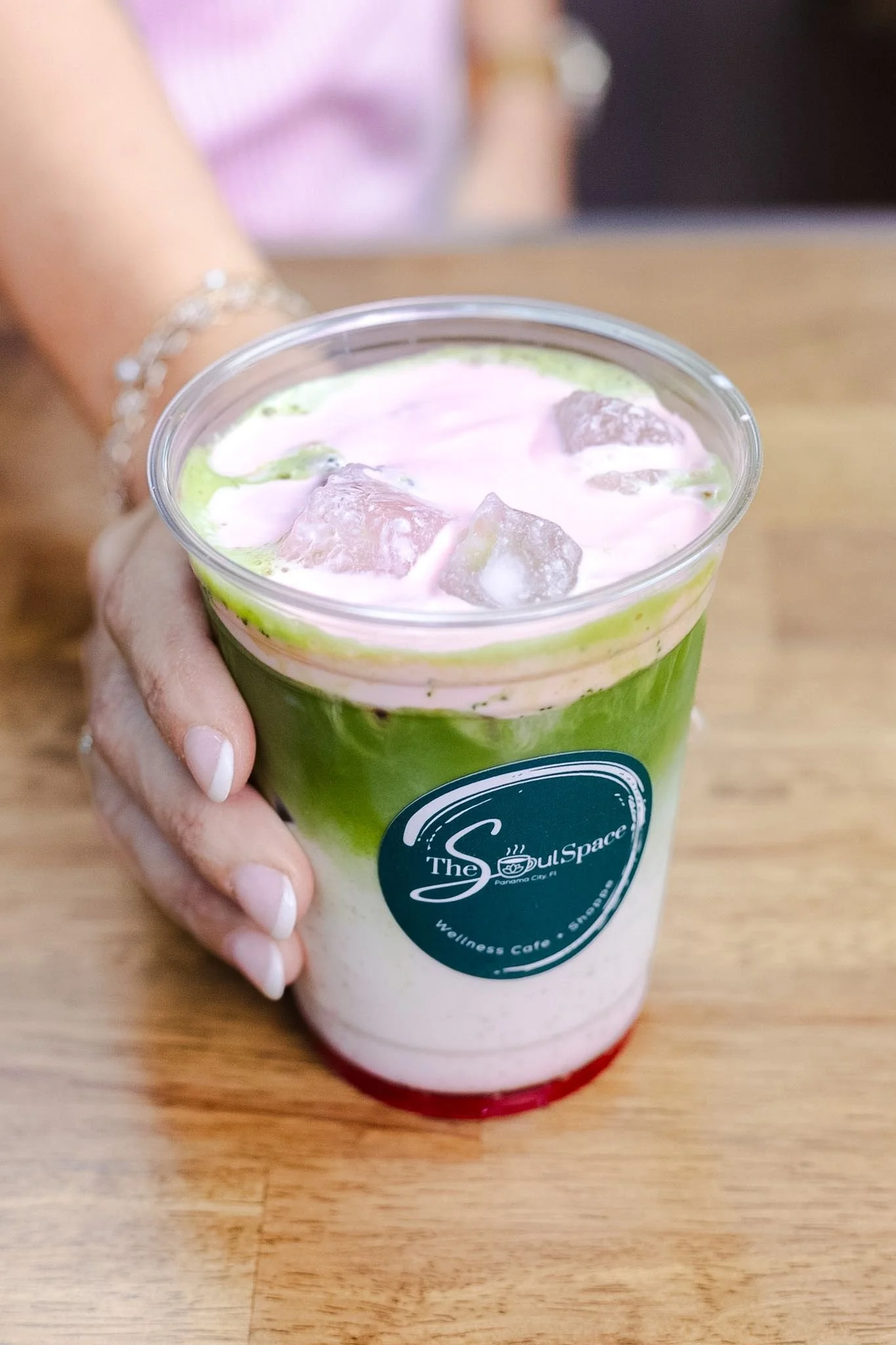 Person holding a colorful layered drink with ice cubes at a coffee shop. Spring menu item: layered iced Matcha.