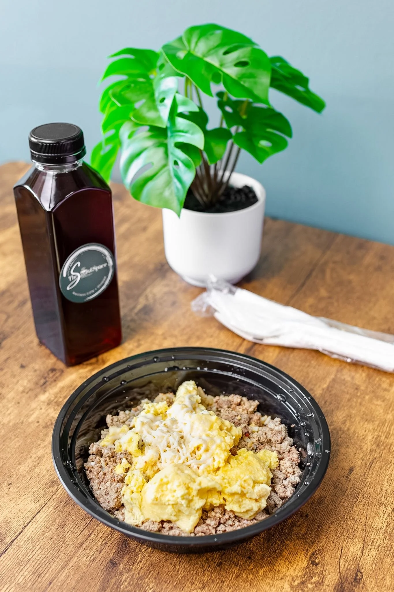 A grab-and-go meal of scrambled eggs and cooked ground beef in a black plastic bowl on a wooden table. Behind the bowl on the table, there's a bottle of cold brew coffee, a white potted plant with green leaves, and a bundle of plastic utensils.