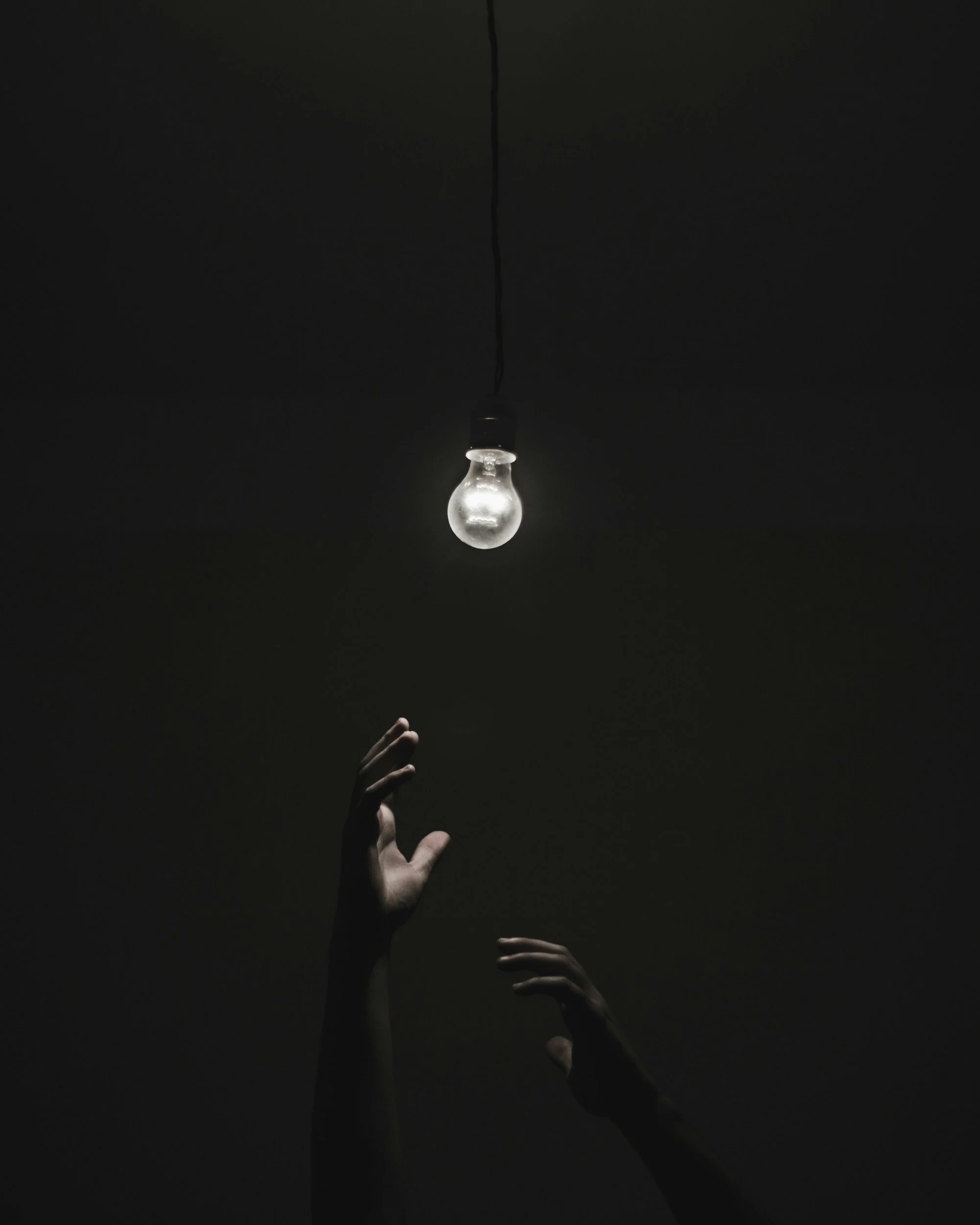 Dark image of a hand reaching toward a hanging light bulb with a black wire against a black background.