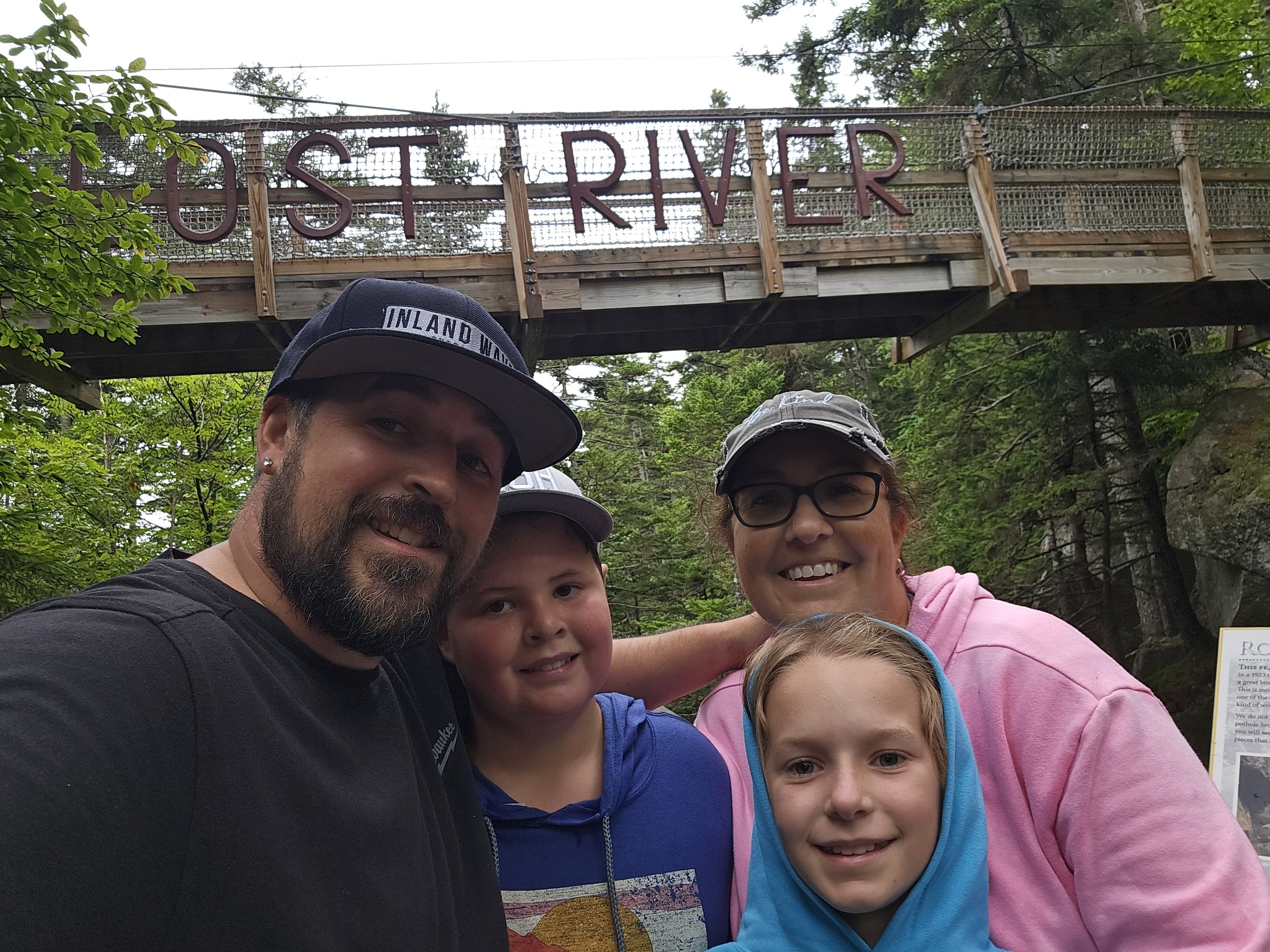 A family of four taking a selfie in front of a wooden bridge with a red sign that reads 'OIST RIVER'. The family consists of a man, woman, and two children, all smiling and dressed casually with outdoor clothing and hats, surrounded by green trees.