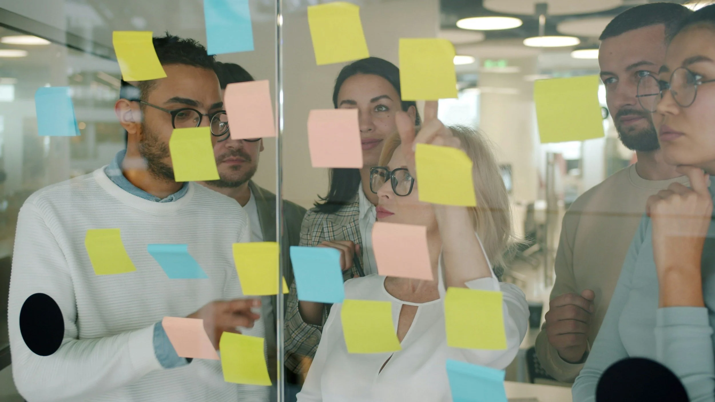 Group of diverse professionals gathered around a glass wall covered with colorful sticky notes, discussing and brainstorming in a modern office.