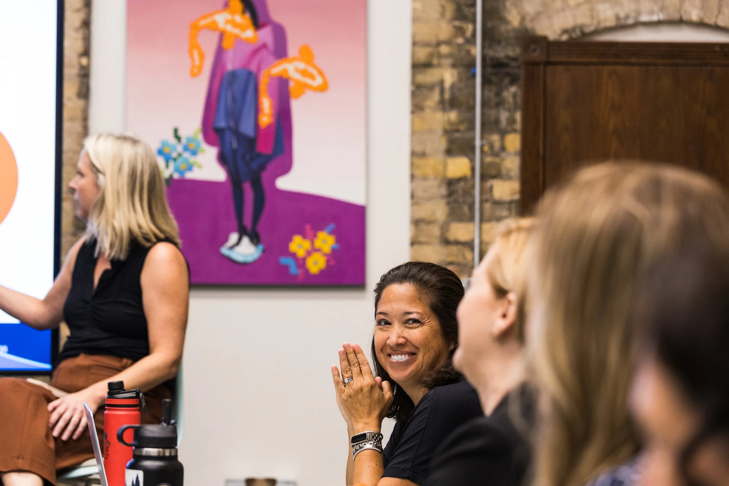 A group of women sitting in a room, smiling and engaged in conversation. One woman is smiling and making a gesture with her hands, while others are listening attentively. A colorful artwork with abstract figures is on the wall behind them.